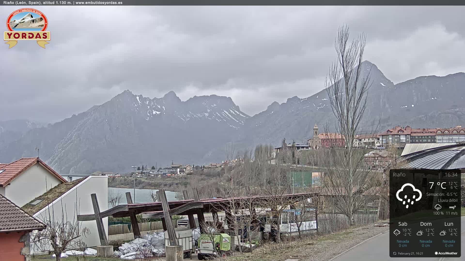 A town nestled in a valley, backed by snow-capped mountains, under a gray, overcast sky with light rain.