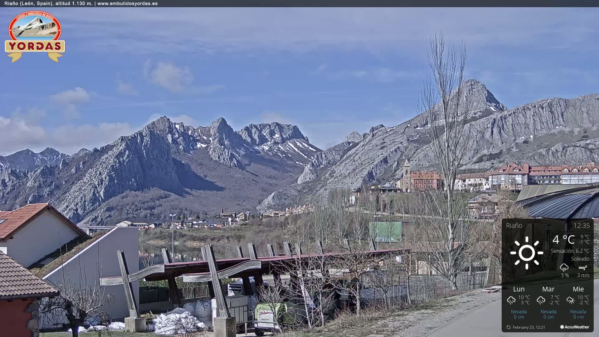 A sunny day reveals a village nestled in a valley, with snow-capped mountains rising dramatically in the background.
