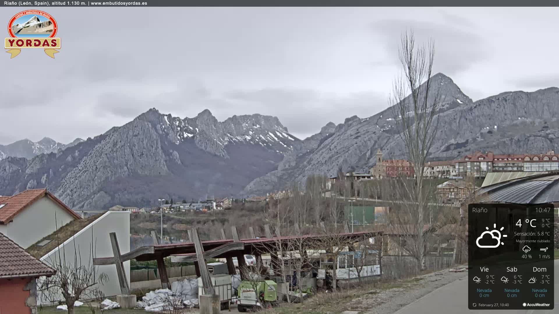 A mostly cloudy day reveals a village nestled in a valley, with snow-capped mountains in the background.