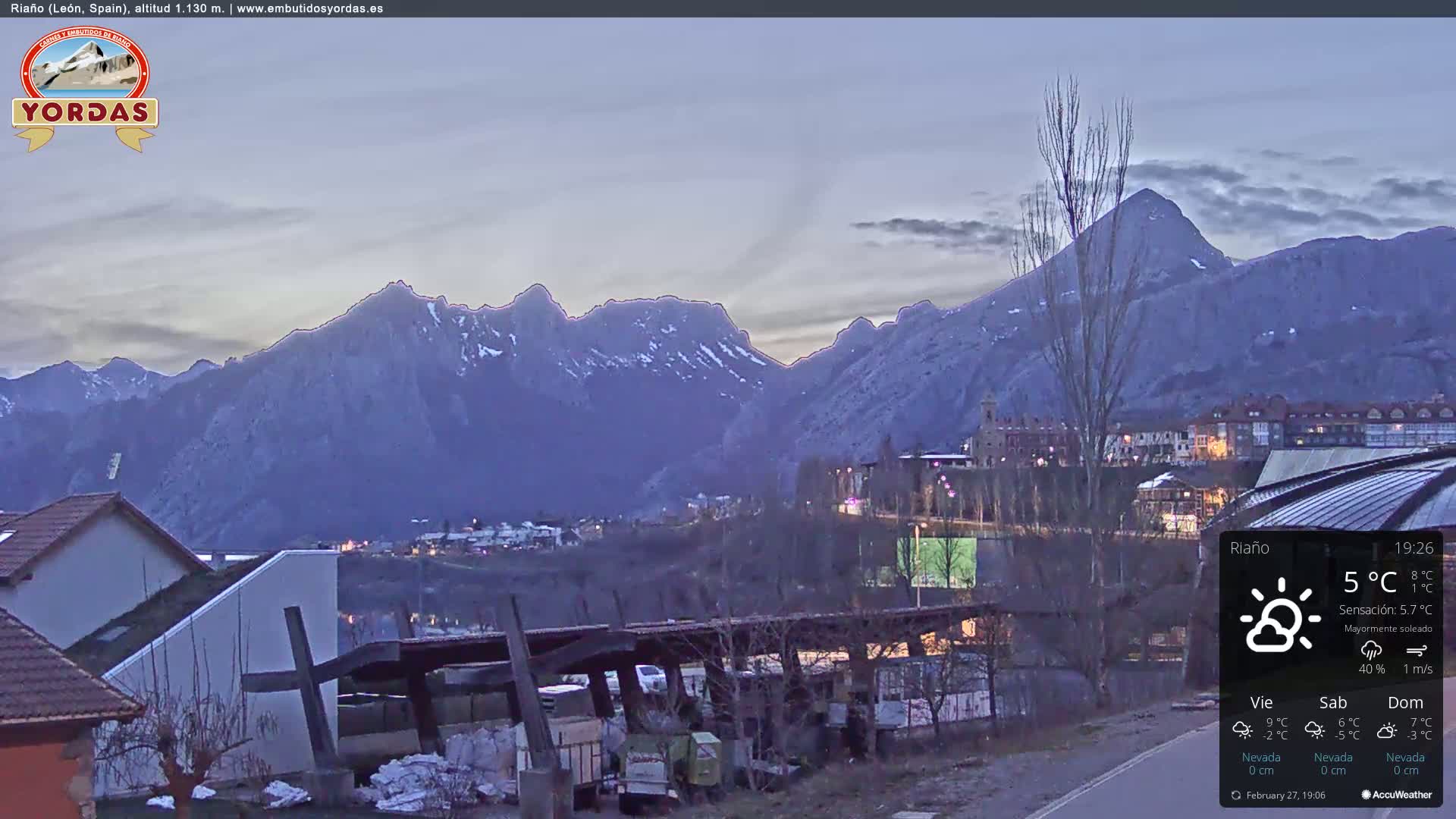 A mountain range dusted with snow is visible at dusk from a hillside town with low-lying buildings and mostly clear skies.