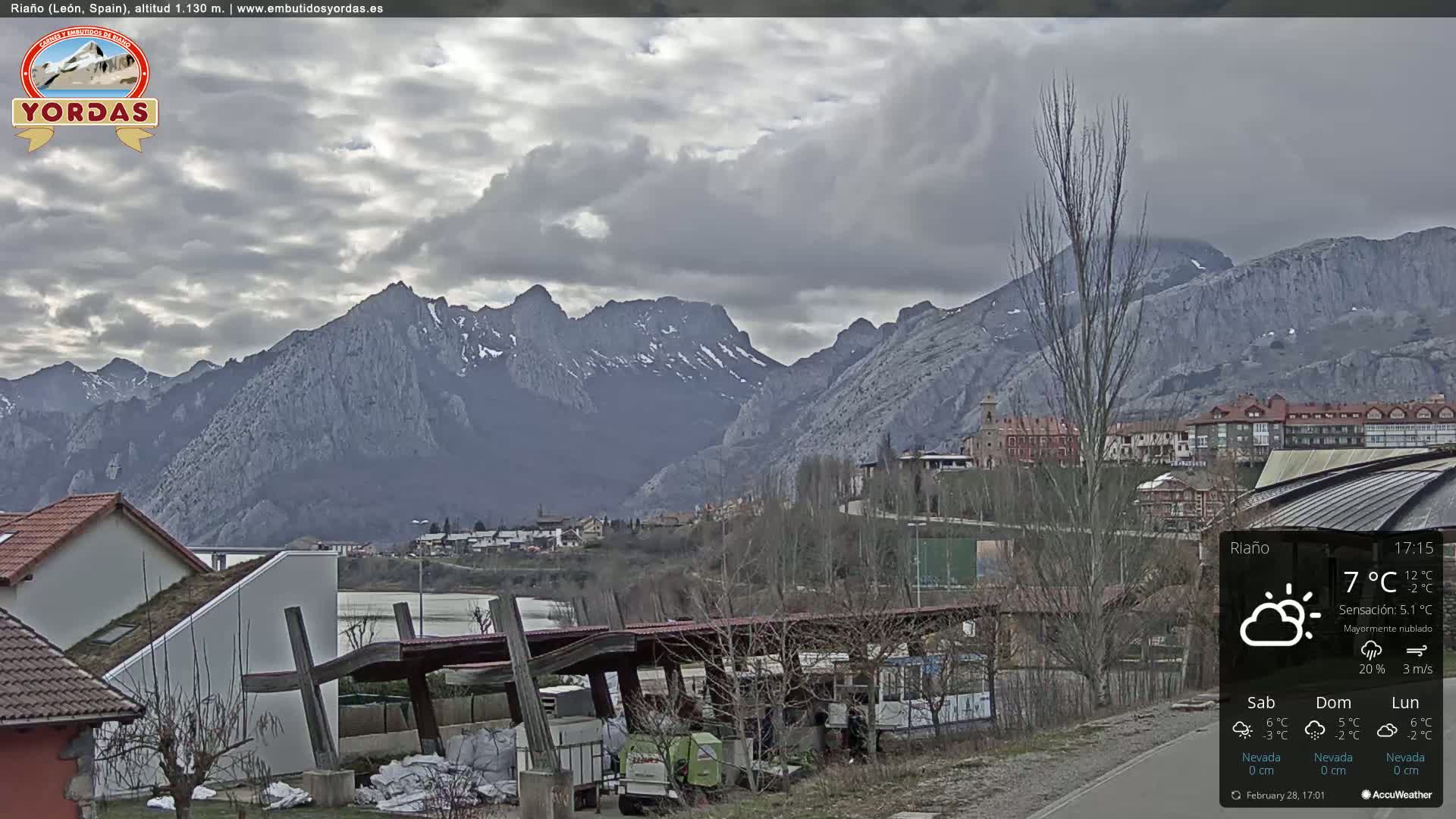 A town nestled at the base of snow-dusted mountains under a mostly cloudy sky.