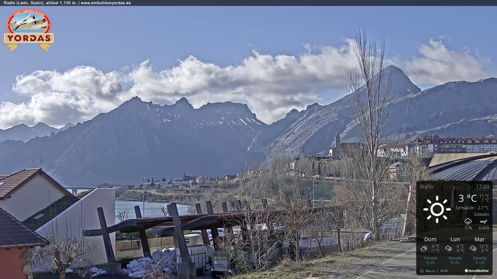 A sunny day reveals a town nestled beside a river, with snow-capped mountains in the background.