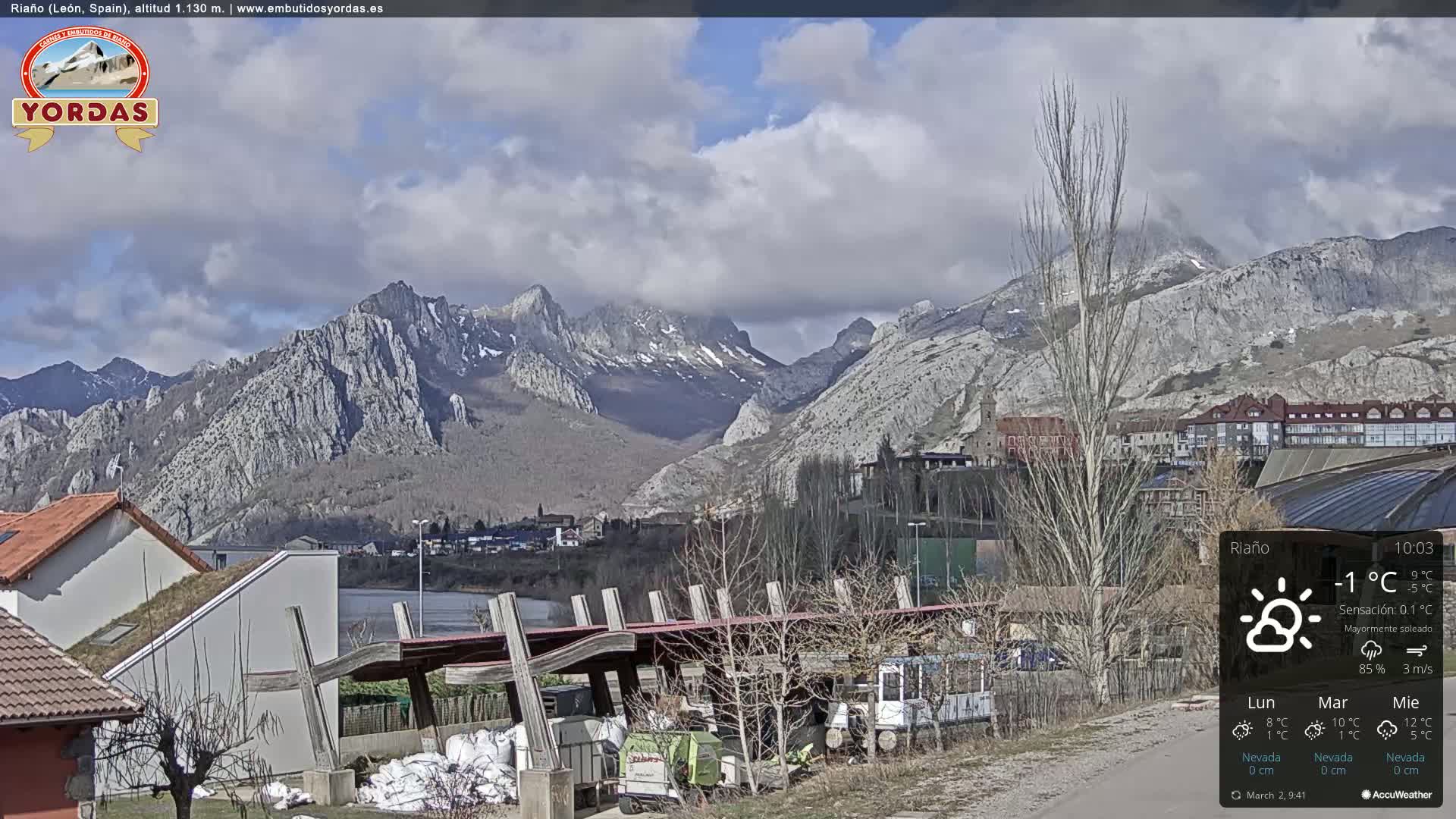 A mostly sunny day shows a town nestled at the base of a mountain range, with some patchy snow visible on the higher peaks.