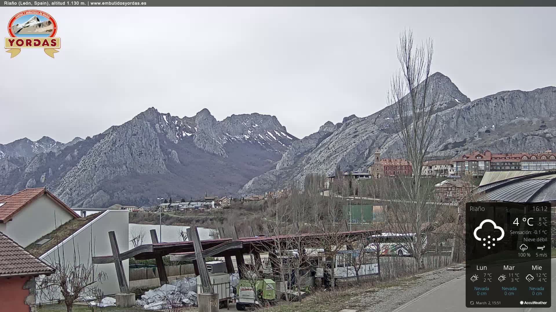 A town nestled in a valley, overlooked by snow-dusted, rocky mountains under a cloudy sky with light snowfall.