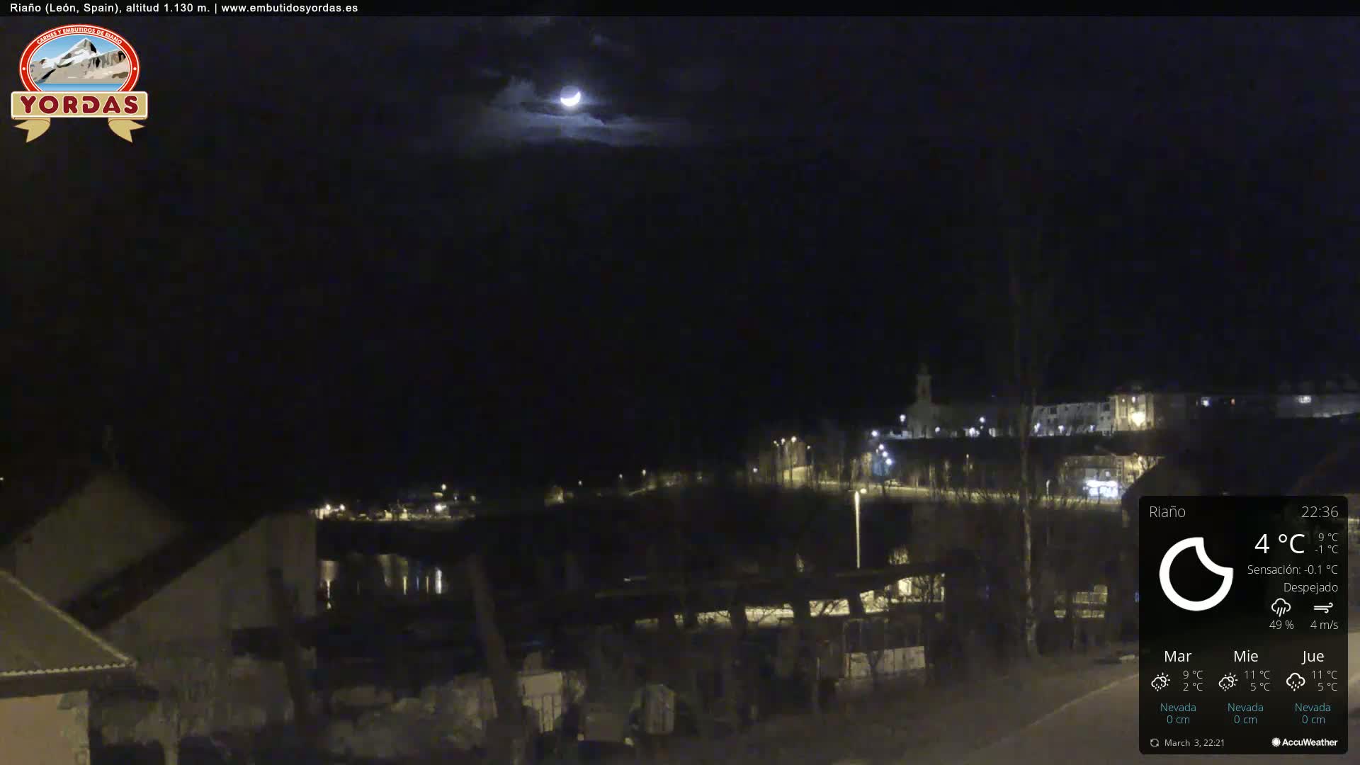 A crescent moon shines in a mostly clear night sky over a dimly lit town.