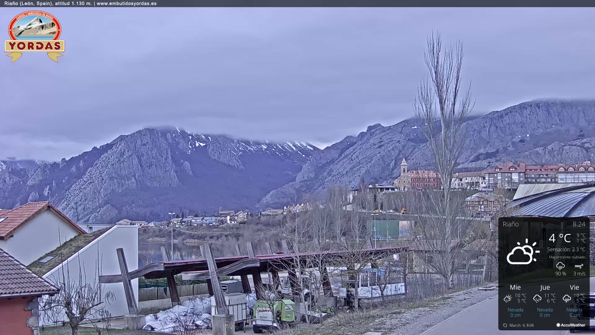 A mostly cloudy view of a town nestled in a valley, with snow-capped mountains in the background.