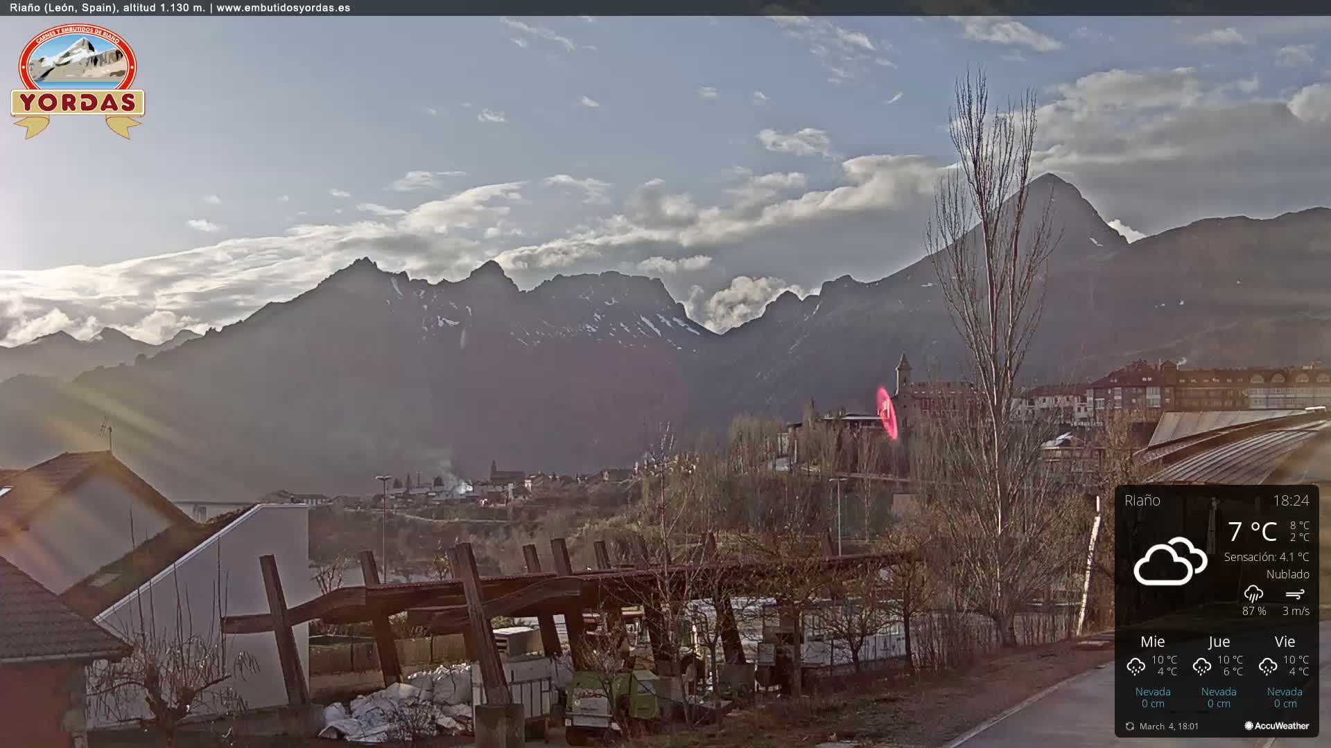 A partly cloudy day reveals a town nestled at the base of a mountain range, with some snow visible on the peaks.