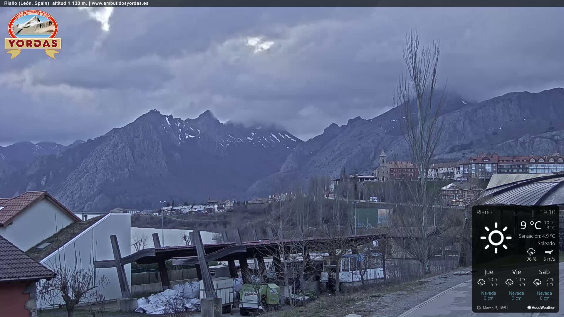 A small town nestled in a valley, overlooked by snow-capped mountains under a cloudy sky.