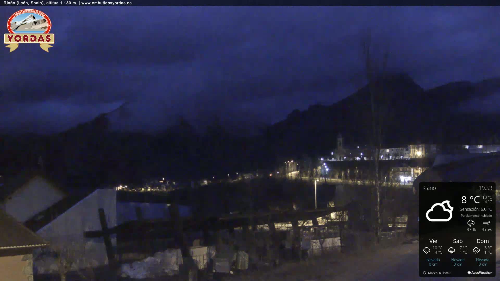 A nighttime view of a small town nestled in a valley between dark mountains under a partly cloudy sky.