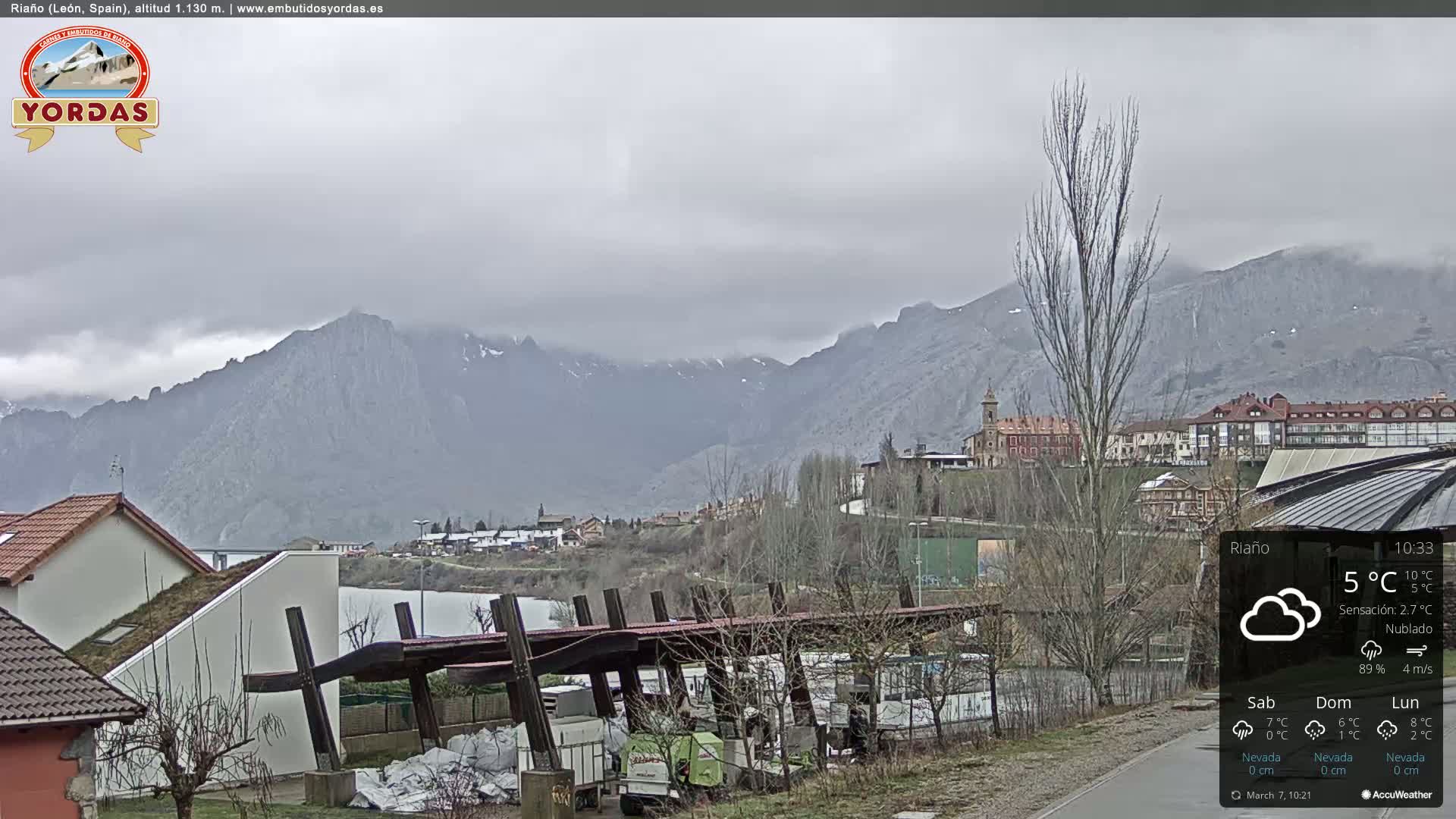 A cloudy day reveals a town nestled at the foot of a mountain range, with a paved path and buildings in the foreground.
