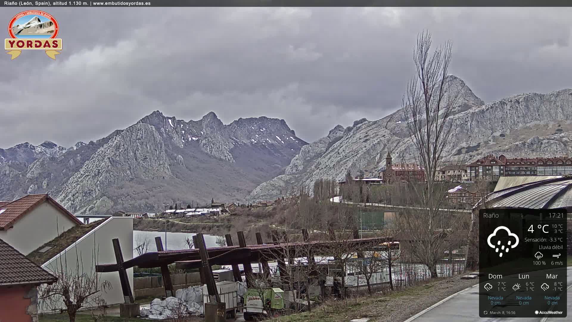 A view of a town nestled in a valley, overlooked by snow-capped mountains under a cloudy, rainy sky.