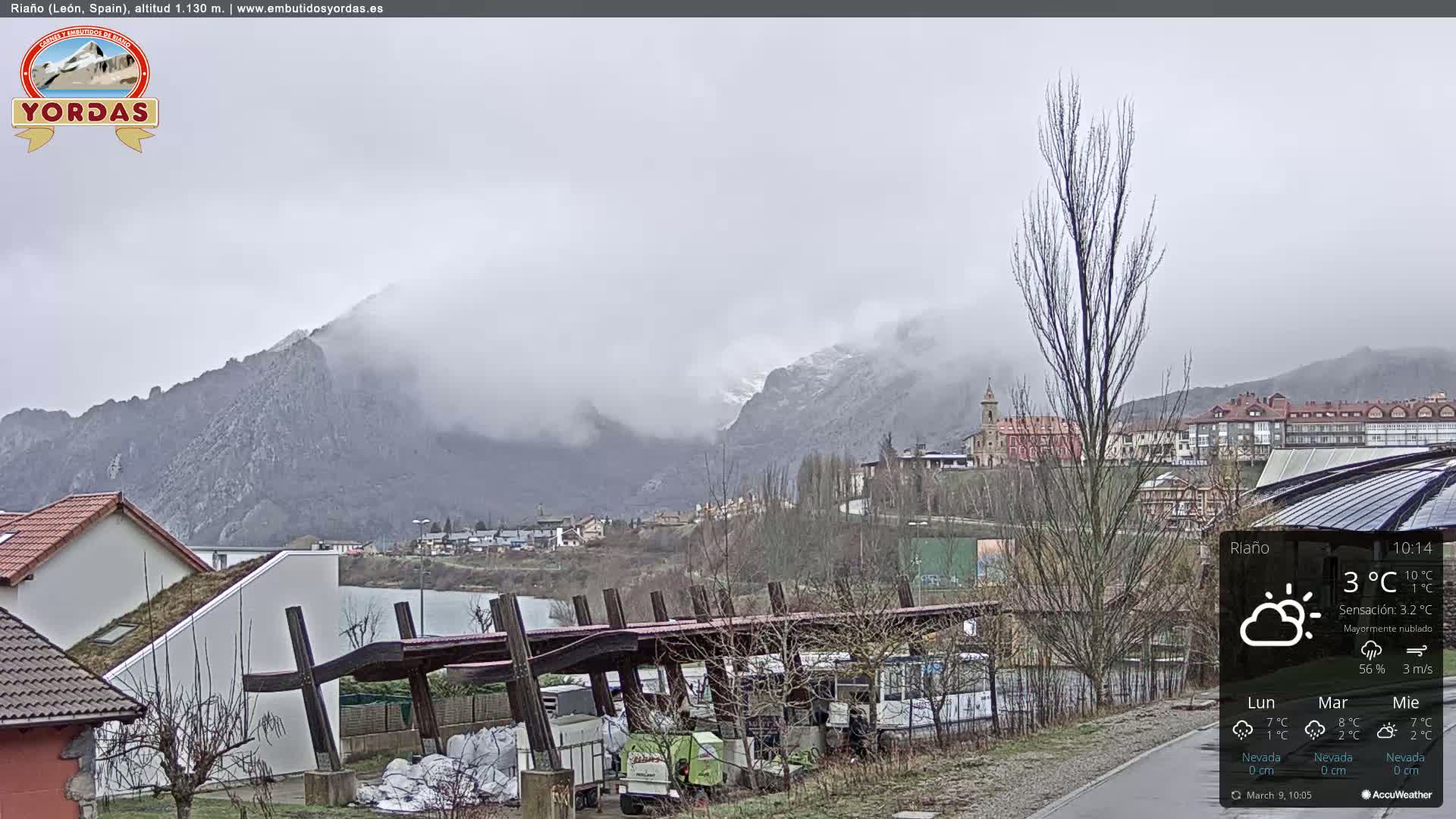 A misty mountain range overlooks a town with buildings and a parking area under a mostly cloudy sky.