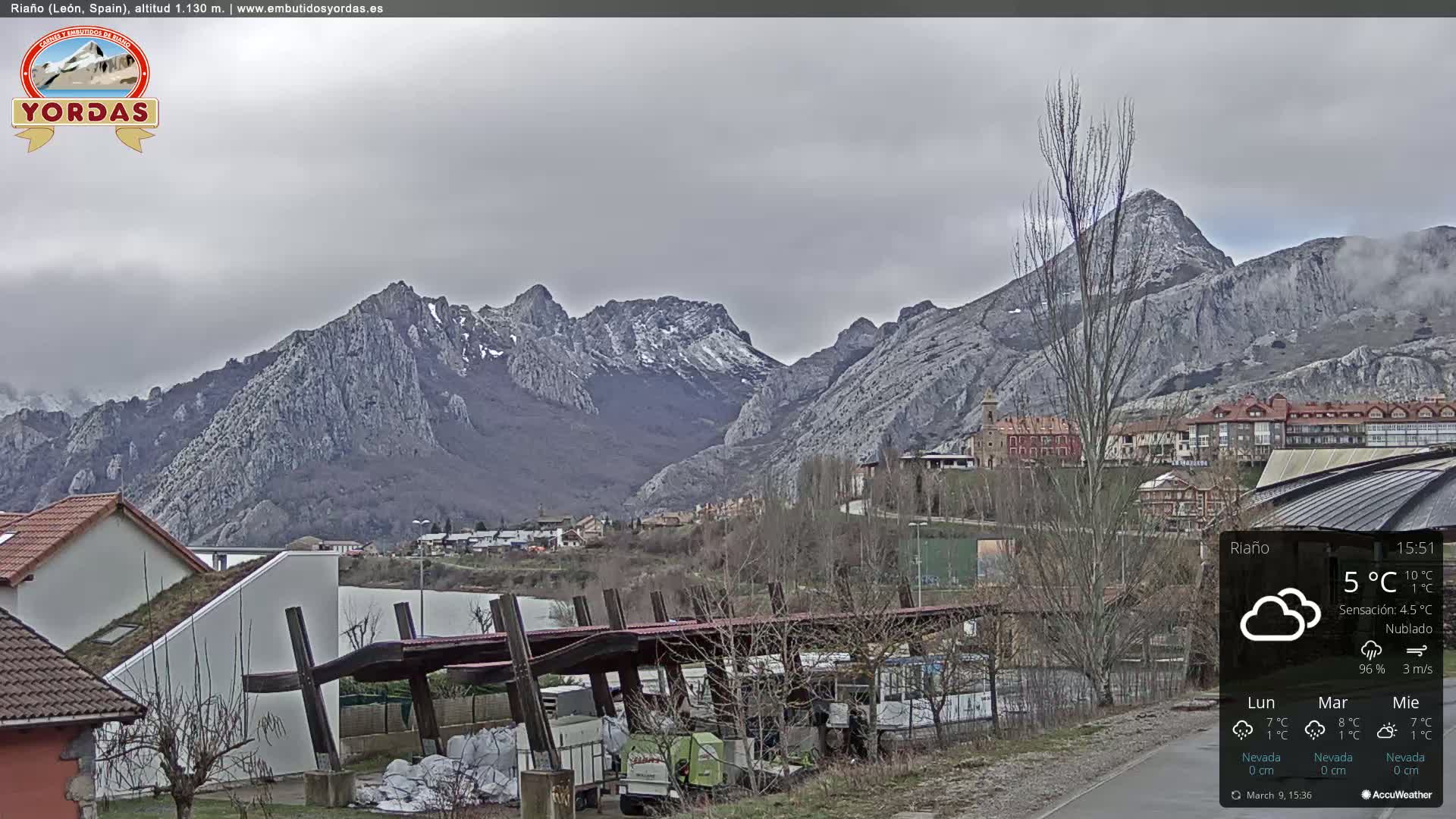 A town nestled in a valley is overlooked by a range of gray, snow-dusted mountains under an overcast sky.