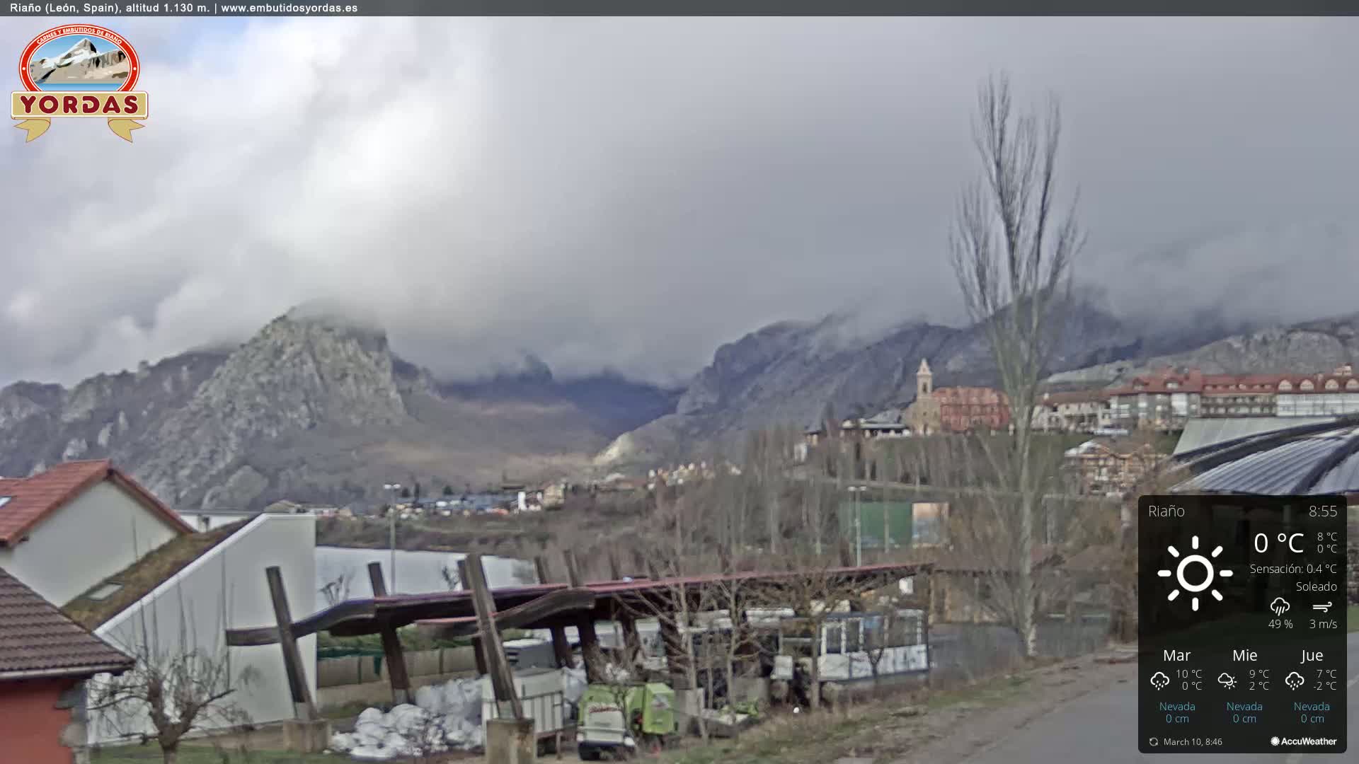 A mountain village is nestled in a valley beneath overcast skies, with bare trees and buildings visible in the foreground.