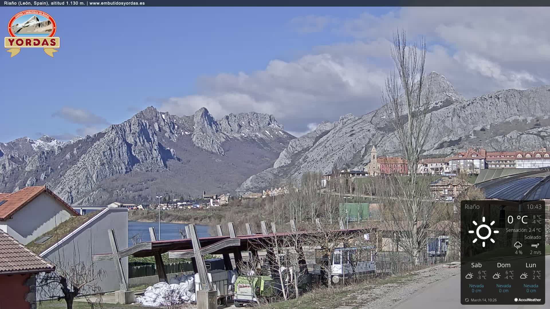 A partly cloudy day reveals a town nestled in a valley, with snow-capped mountains in the background and a body of water visible in the foreground.