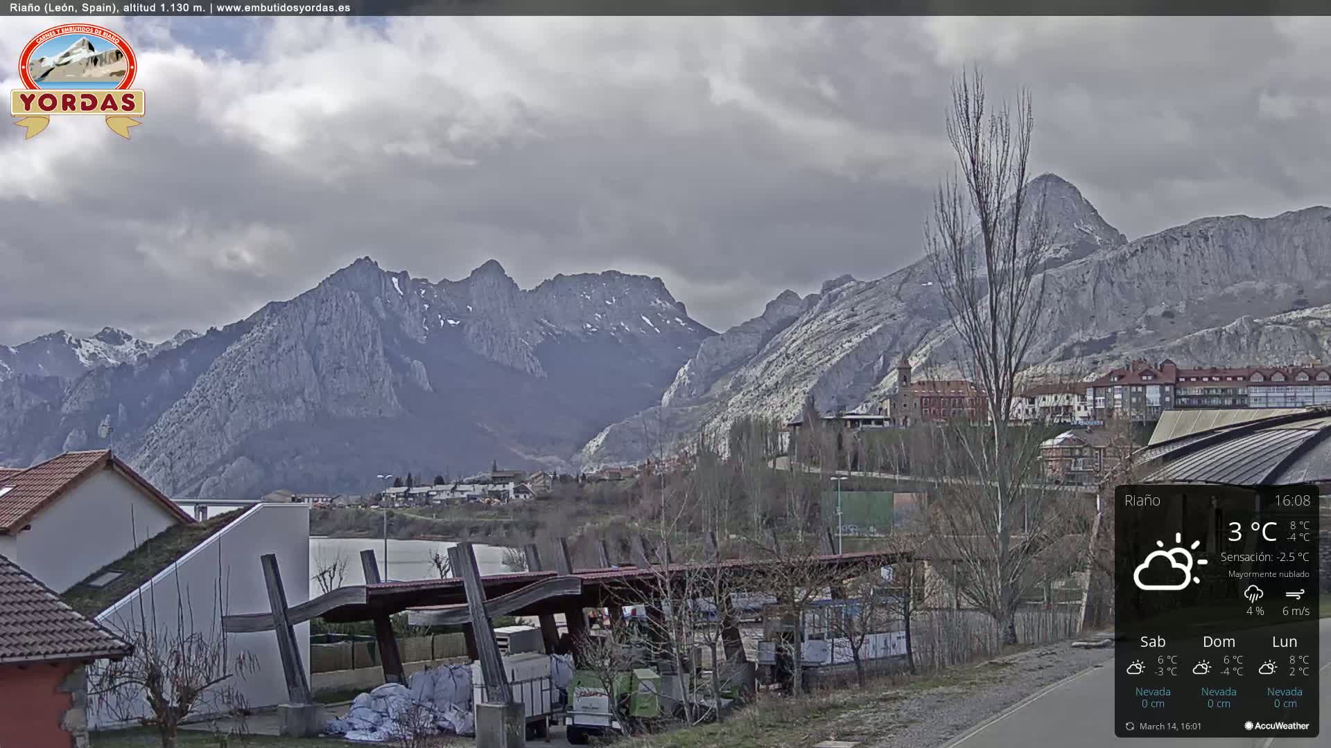 A mostly cloudy day reveals a village nestled at the base of a large, imposing mountain range.