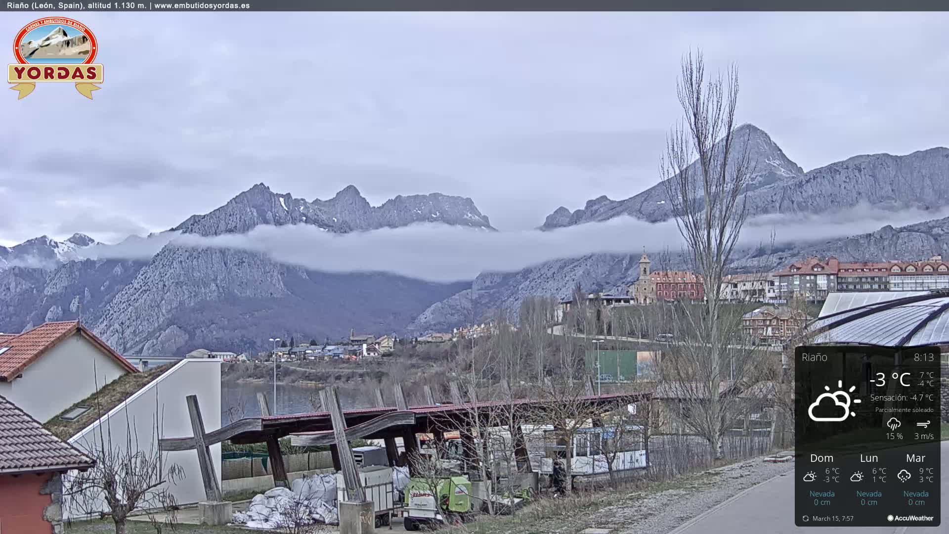 A partly cloudy day reveals a mountain range partially obscured by low clouds, overlooking a town with buildings and a lake.