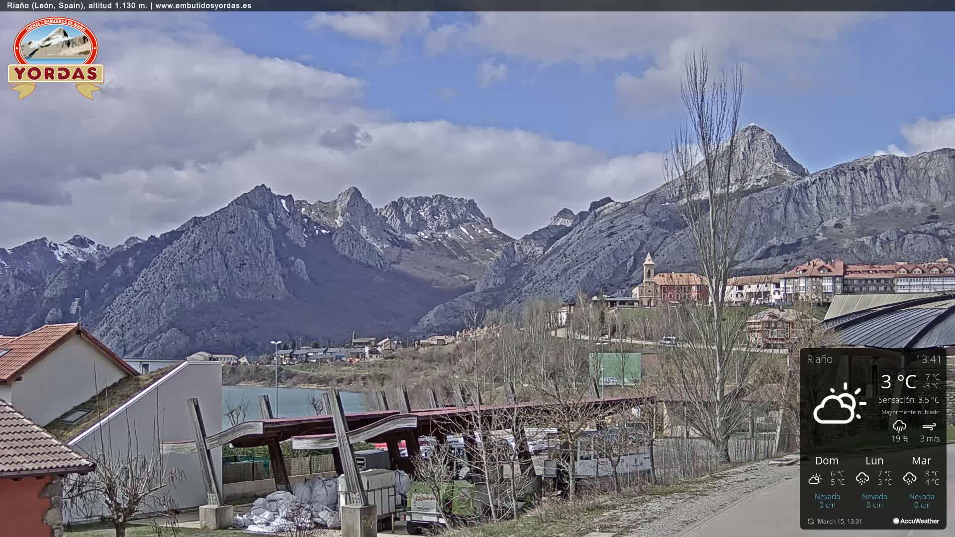 A partly cloudy day reveals a mountain range with some snow, overlooking a town with buildings nestled amongst bare trees.