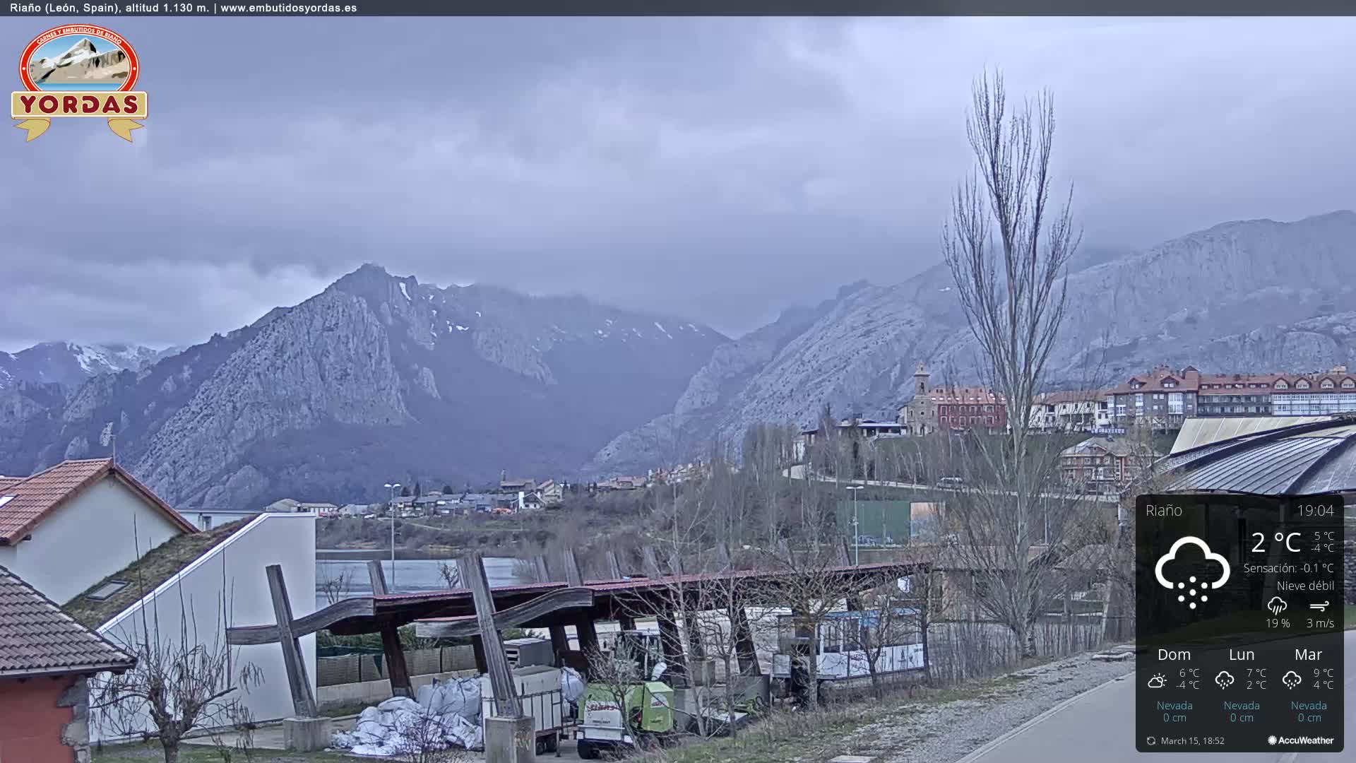 A snow-dusted mountain range overlooks a town situated beside a body of water, under a cloudy sky.