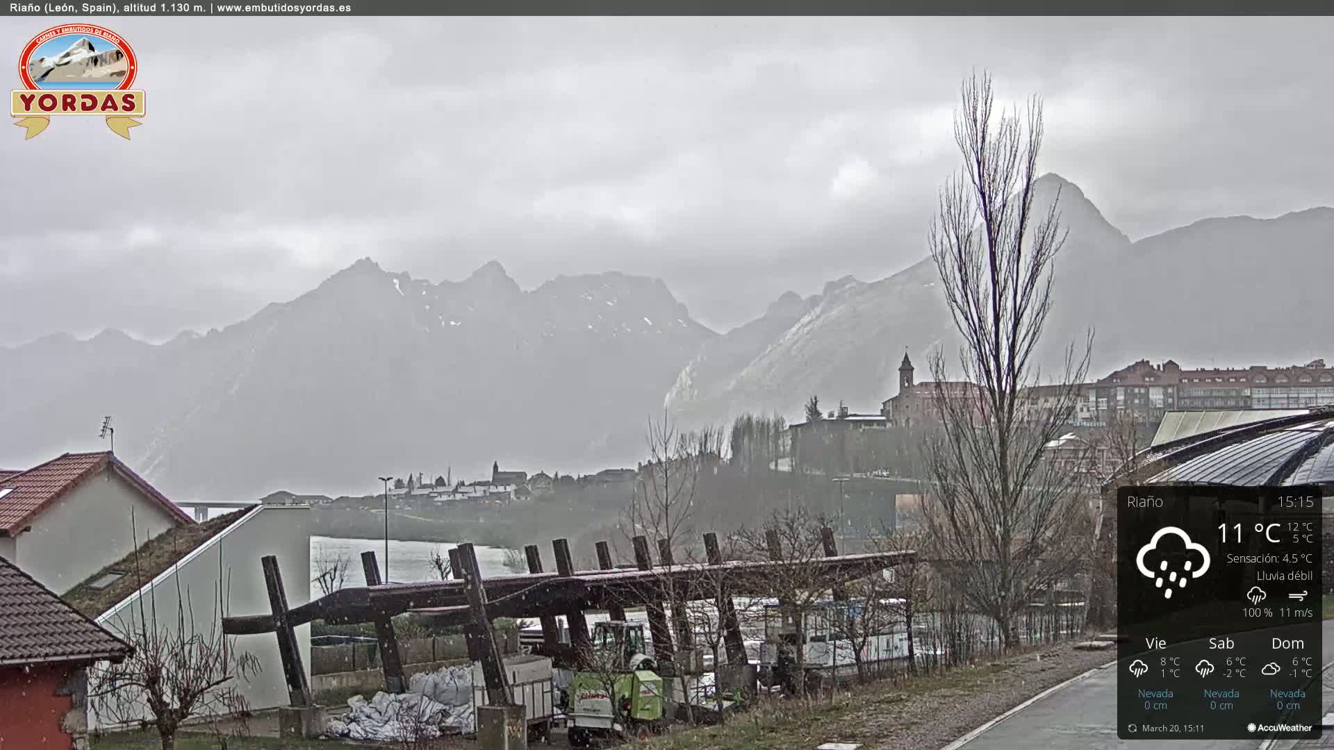 A light rain falls on a town nestled beside a lake and mountains under a gray sky.