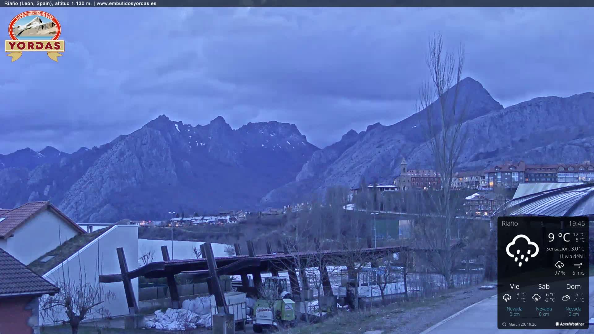 A mountain range is visible in the distance under an overcast sky with light rain, overlooking a town with buildings and a few trees, near a parking area with machinery.