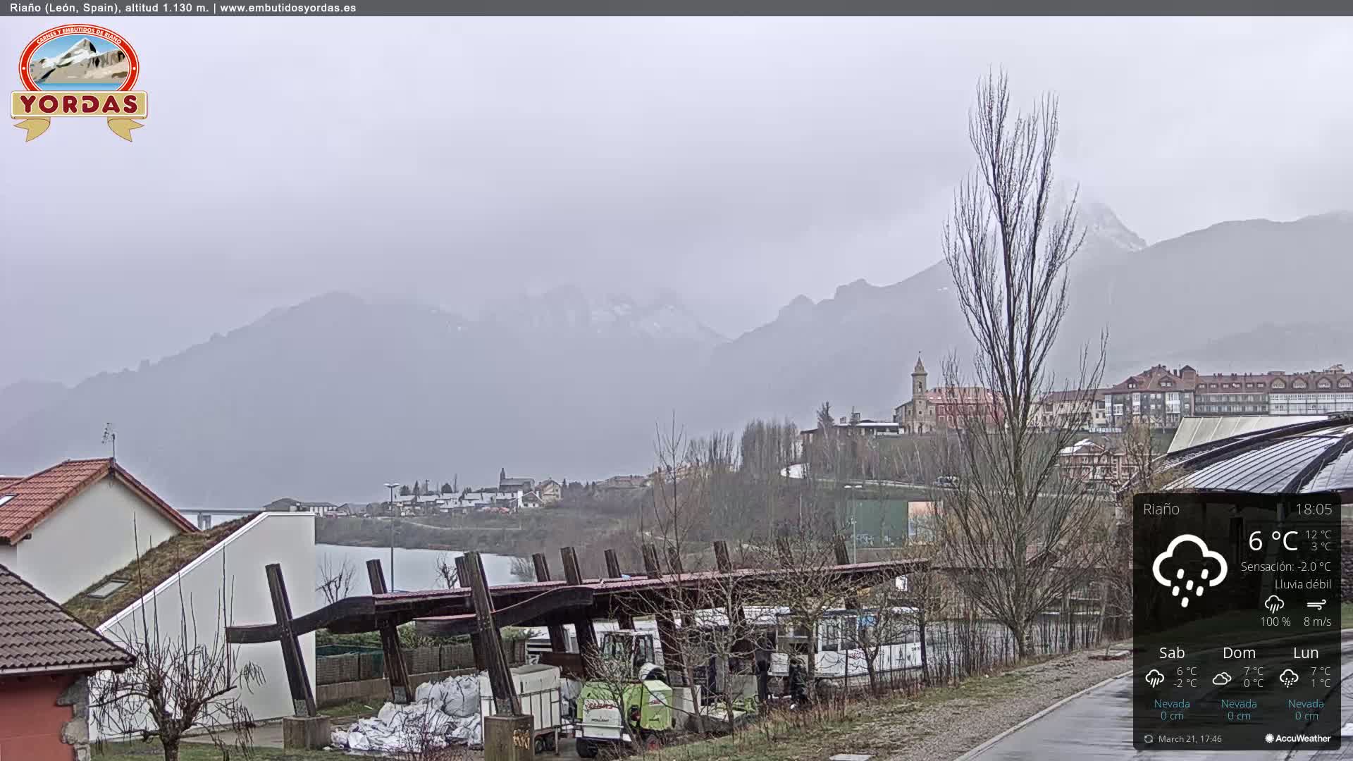 A view of a town nestled in a valley between snow-capped mountains under a gray, overcast sky with light rain.