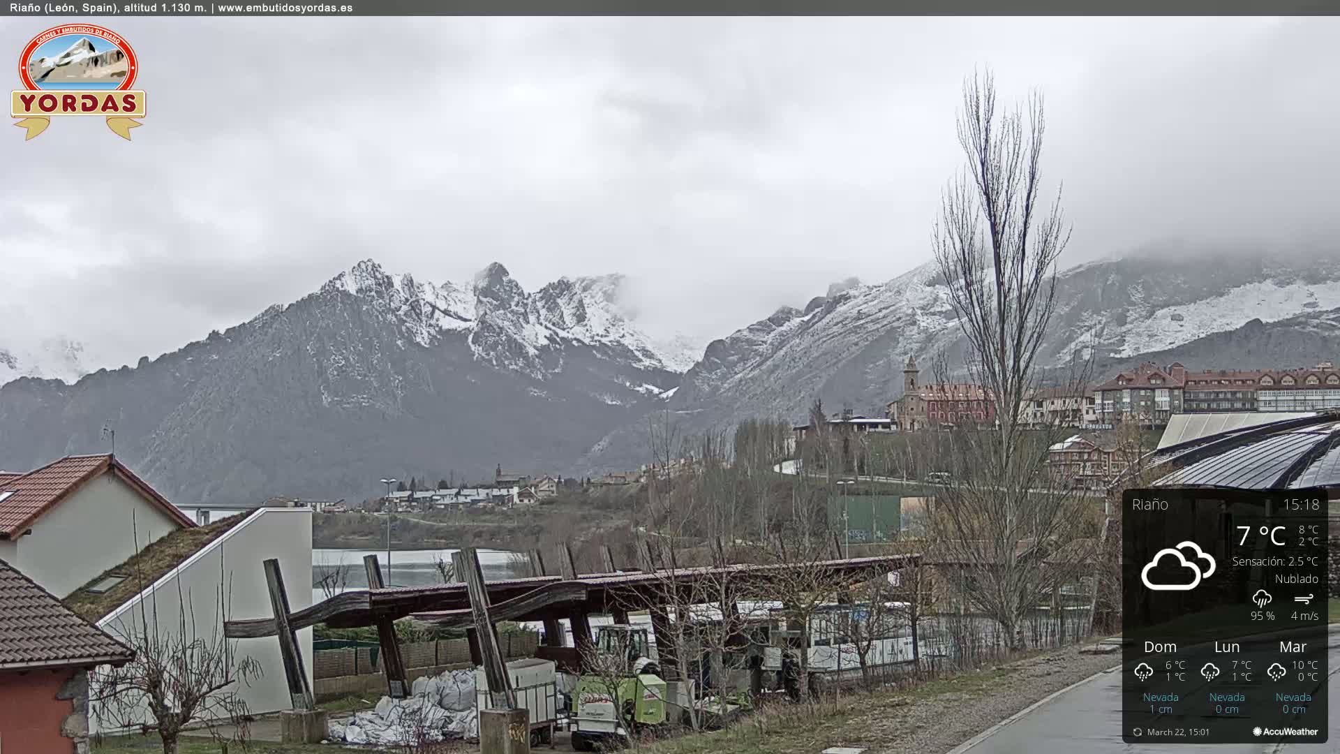 A cloudy day reveals a snow-capped mountain range behind a small town nestled in a valley, with some buildings and vehicles visible in the foreground.