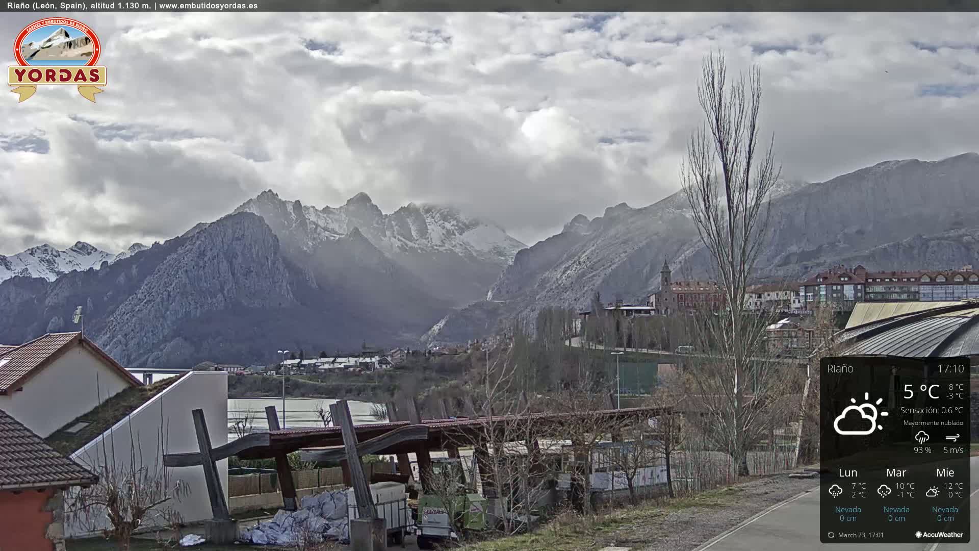 A partially cloudy day reveals snow-capped mountains in the distance behind a town with modern and older buildings, some houses and a partially constructed structure in the foreground.