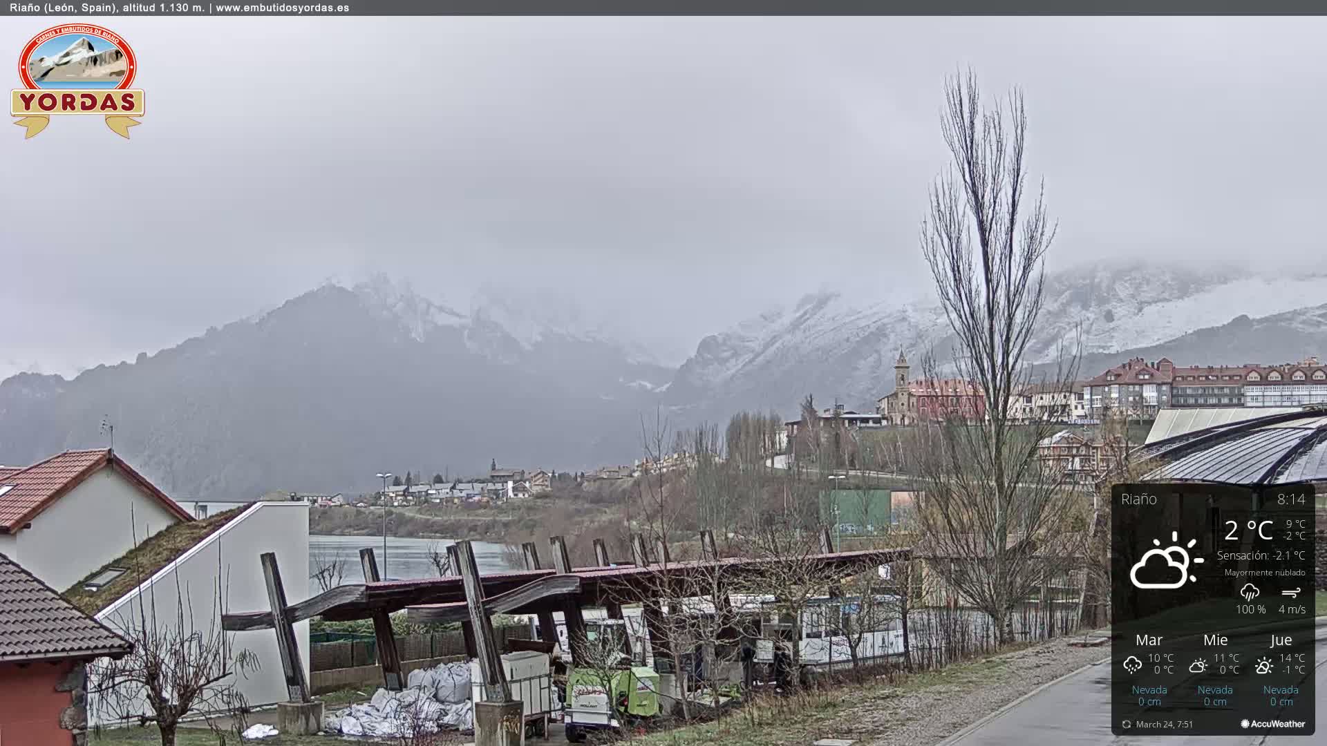A mostly cloudy, gray day reveals a snow-capped mountain range in the distance, overlooking a town with buildings and a body of water in the foreground.
