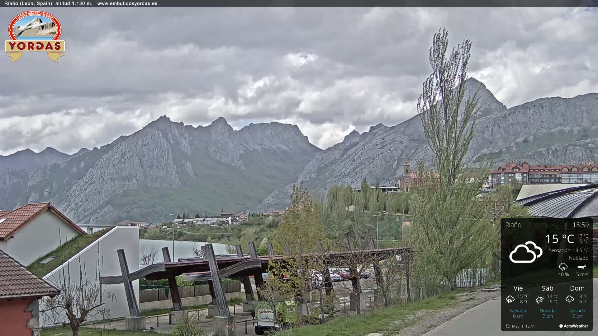 A cloudy day reveals a mountain range behind a town with some buildings and a partially covered parking area.