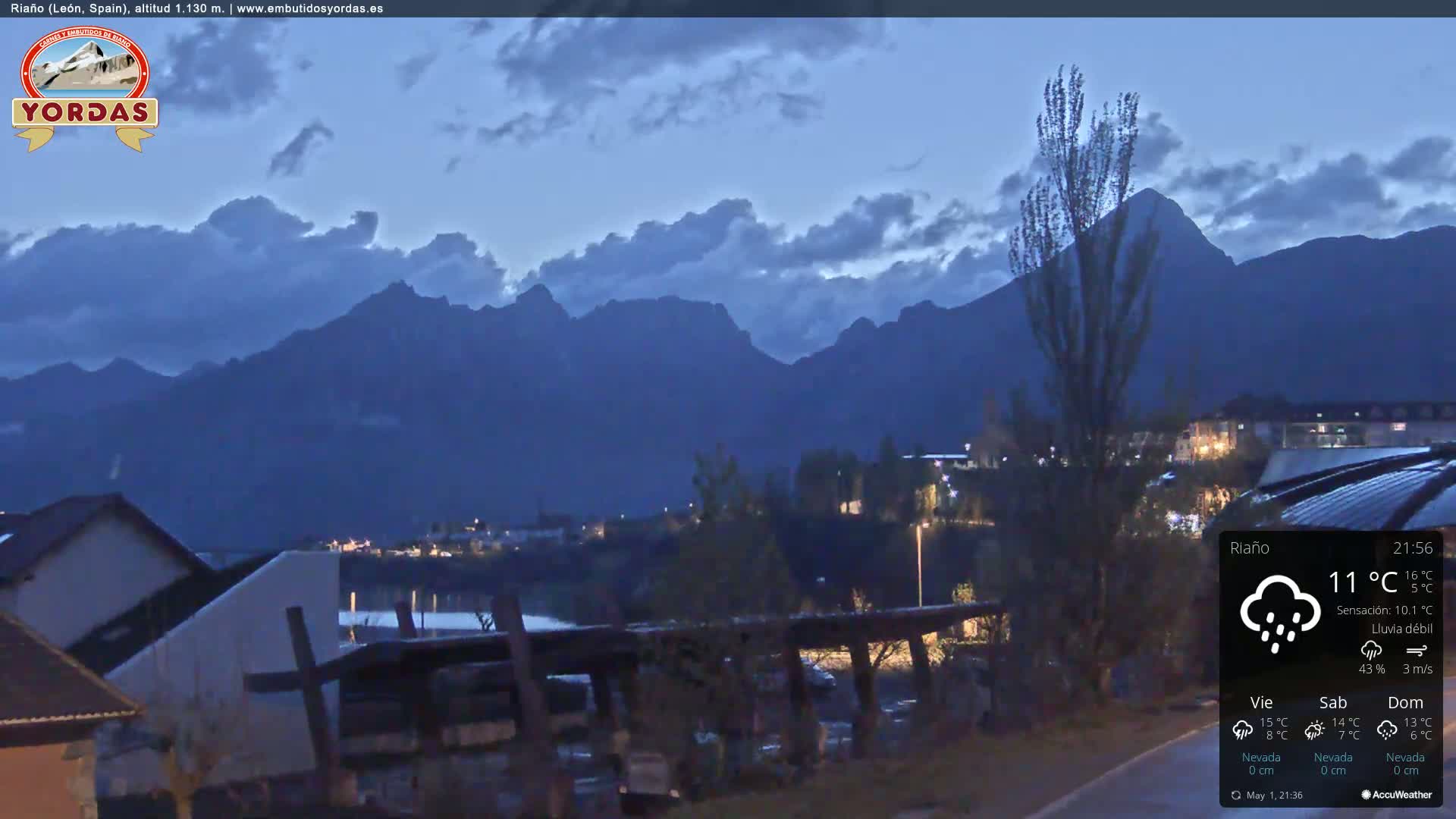 A nighttime view of a town nestled against a mountain range under a partly cloudy sky with light rain.