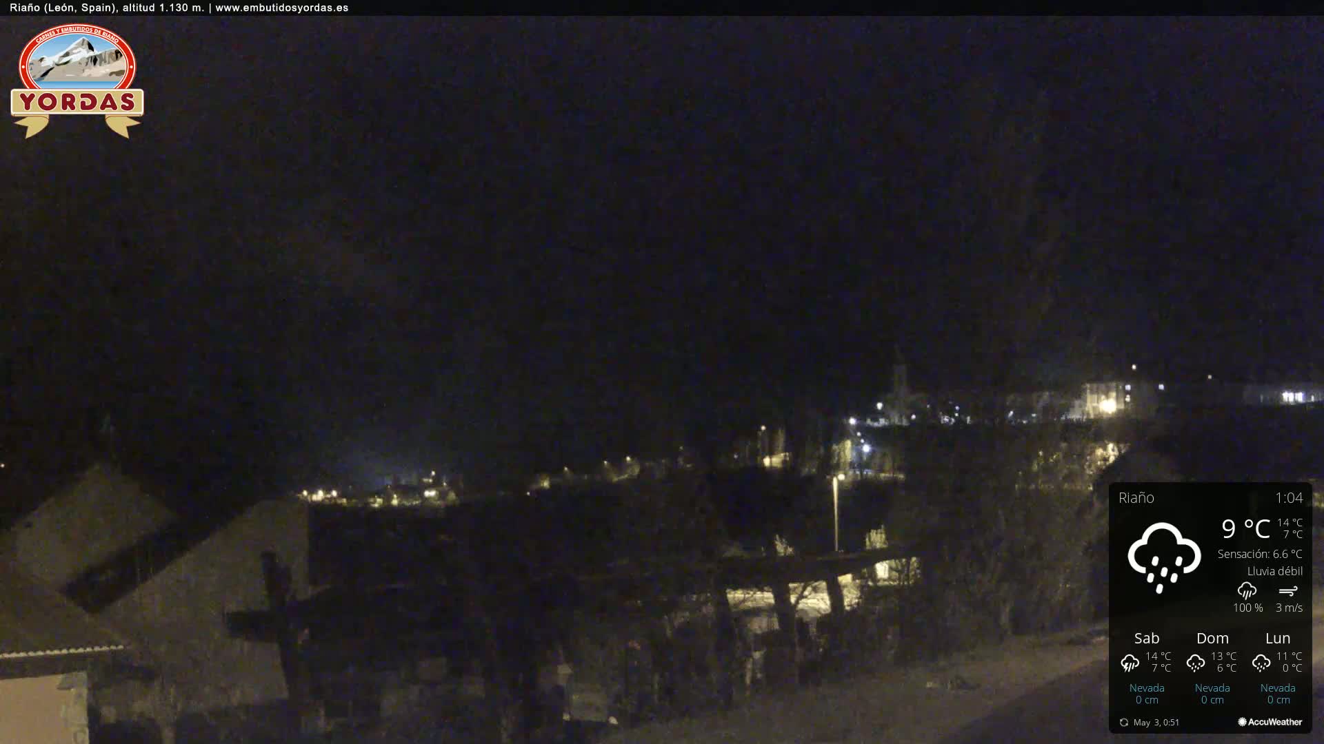 A nighttime view of a small town, lightly illuminated, under a dark sky with light rain.