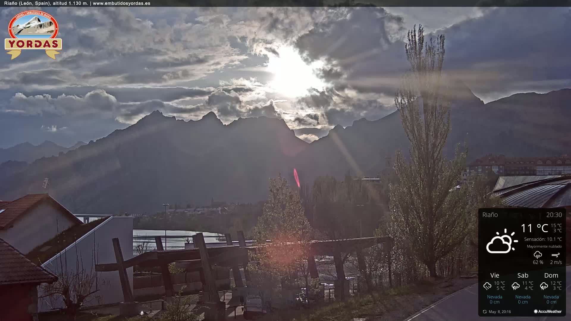 A mostly cloudy sunset illuminates a mountain range behind a town with houses and trees, viewed from an elevated perspective.