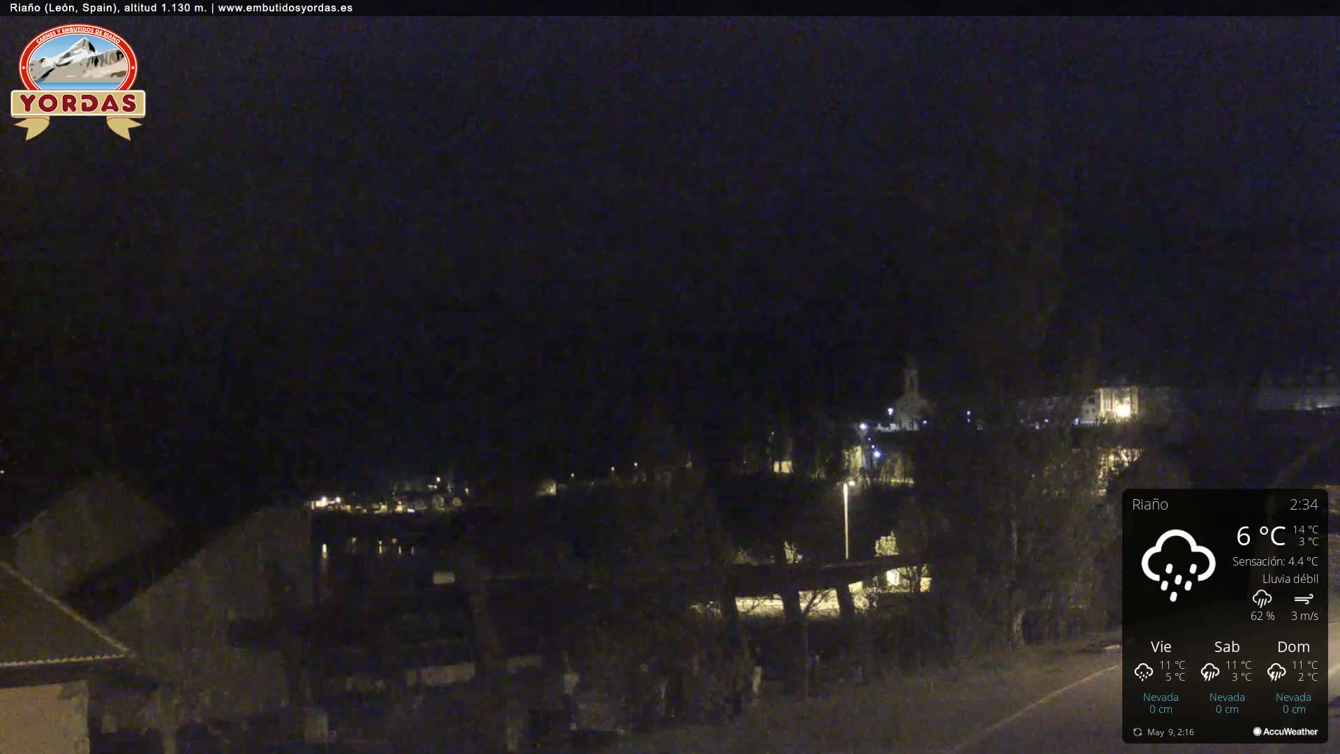 A nighttime view of a small town under light rain, with dimly lit buildings and sparsely populated streets.