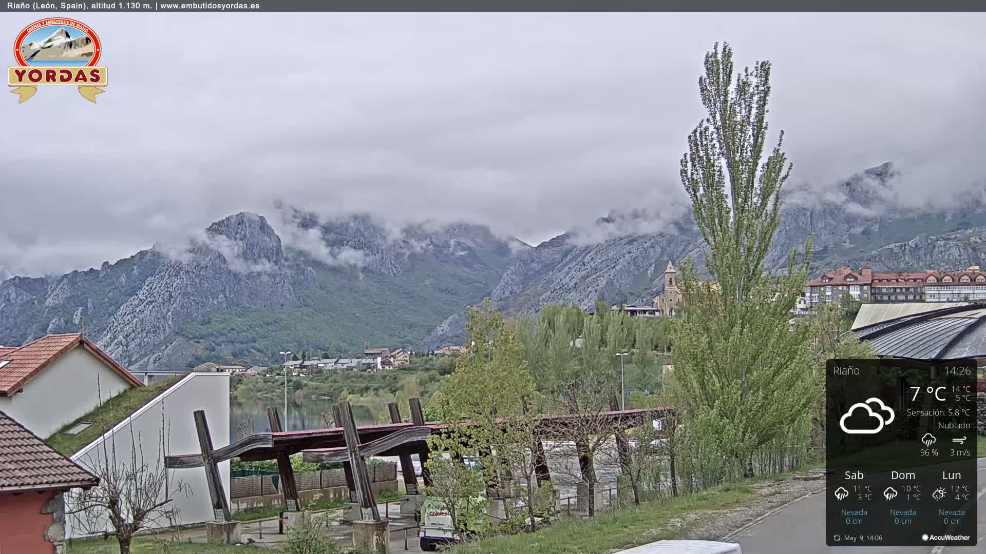 A cloudy day reveals a town nestled at the base of misty mountains, a body of water visible in the foreground.