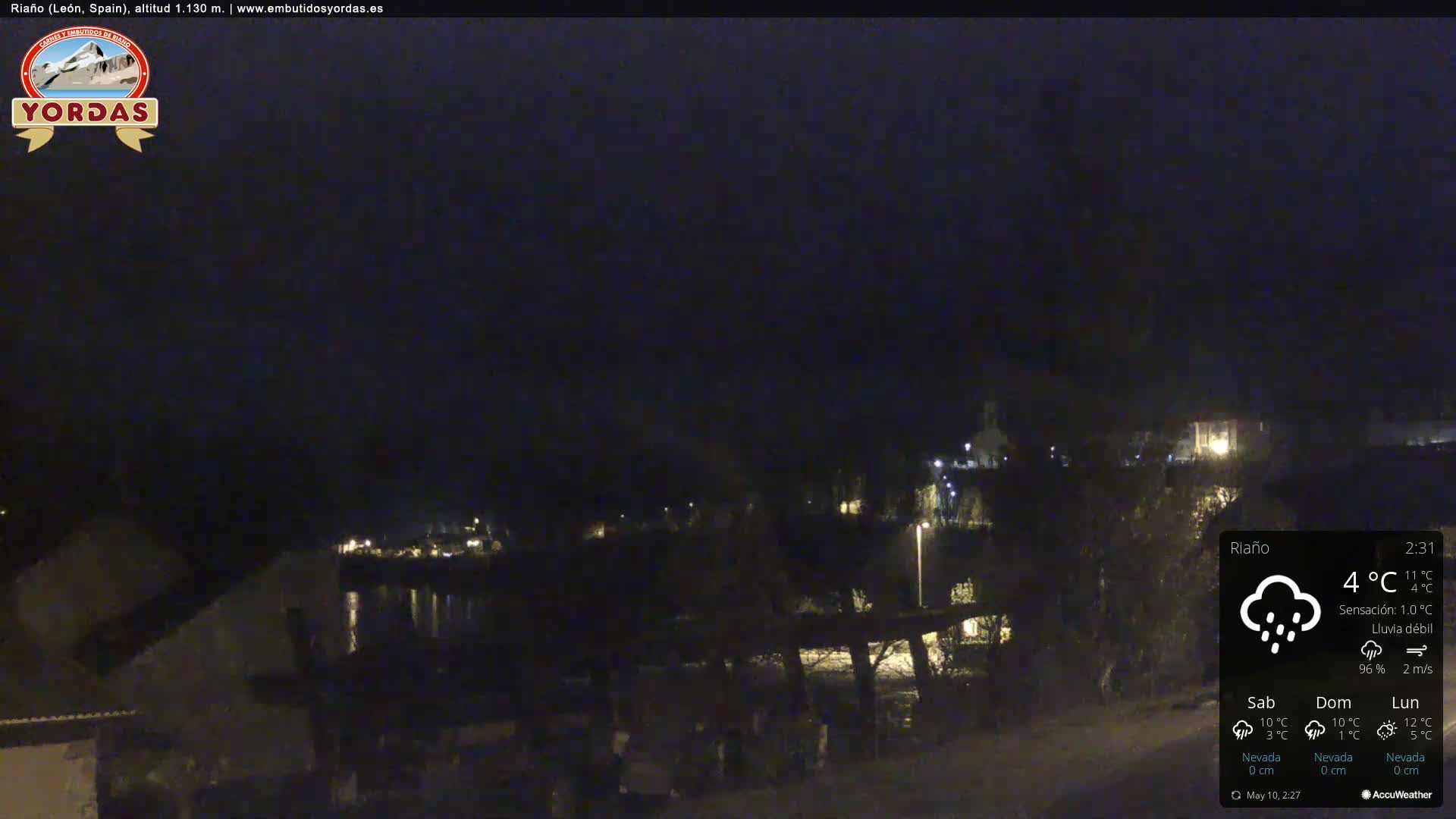 A nighttime view of a dimly lit town beside a body of water under a dark sky with light rain.