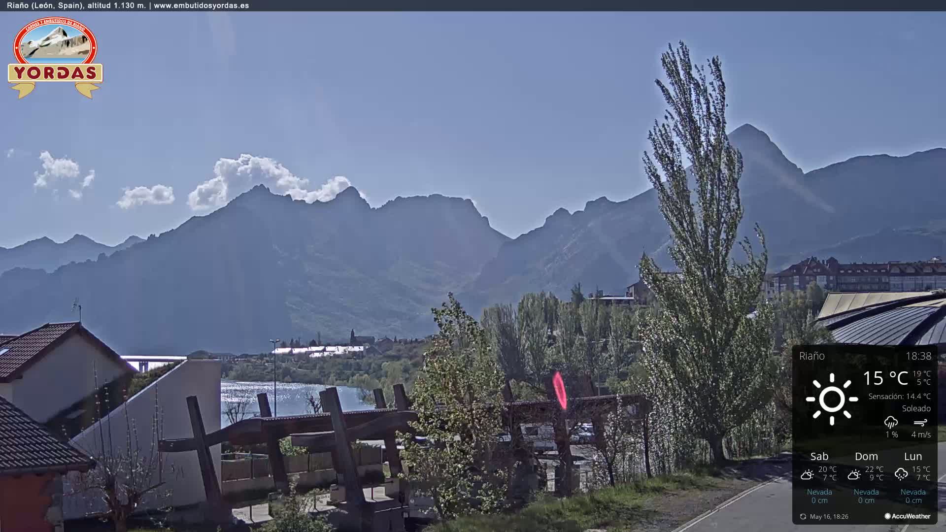 A sunny day reveals a view of a lake, buildings, and trees nestled at the foot of a mountain range under a mostly clear sky.