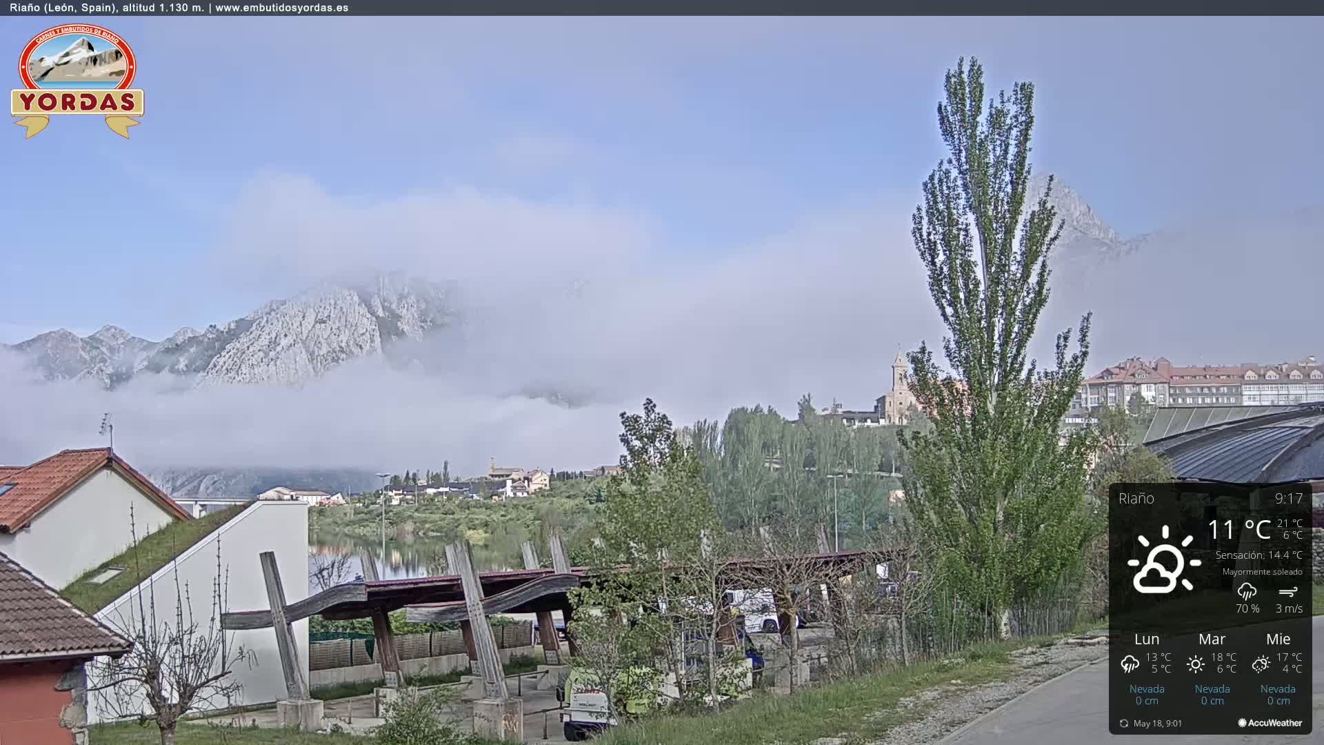 A mostly sunny day with a low fog partially obscuring mountains in the distance, overlooking a town with a lake and modern and traditional buildings.
