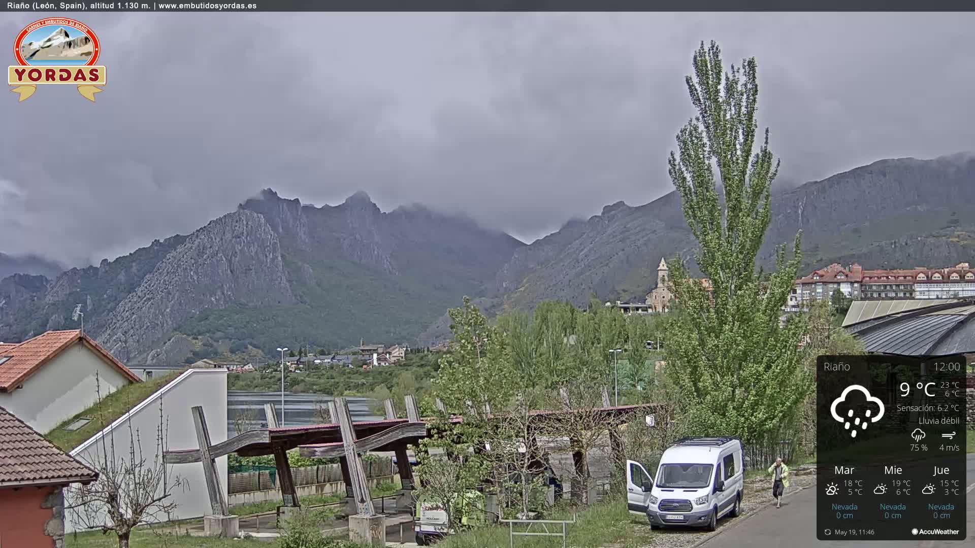 A rain-swept view of a town nestled in a valley, backed by imposing mountains under a cloudy sky.