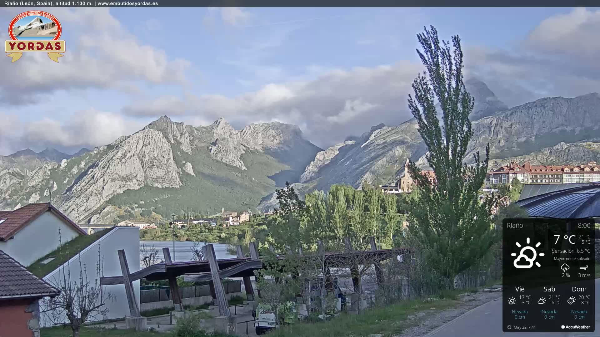 A partly cloudy day reveals a town nestled in a valley, backed by imposing, grey, rocky mountains.