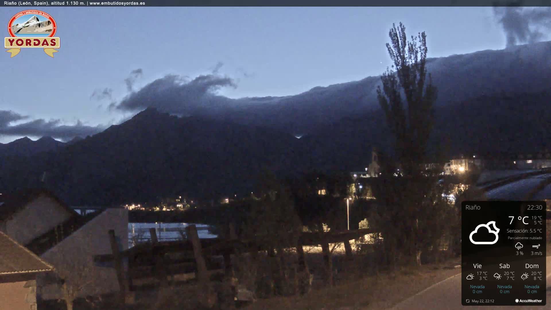 A nighttime view of a town nestled at the base of dark mountains under a partly cloudy sky.
