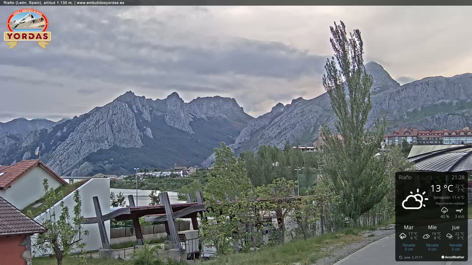 A mostly cloudy view shows a town nestled at the foot of a large, grey mountain range.