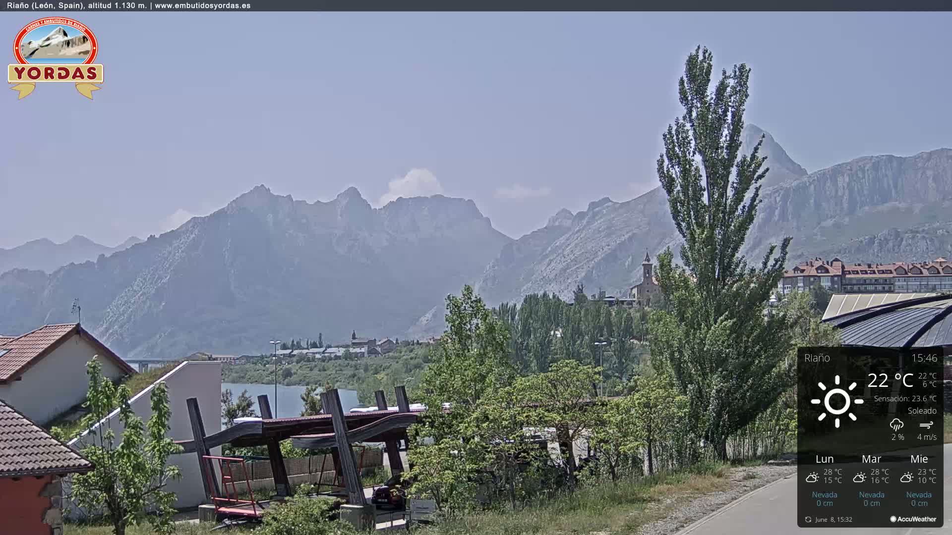 A sunny view of a town nestled in a valley surrounded by large, hazy mountains, a calm lake is visible in the foreground.