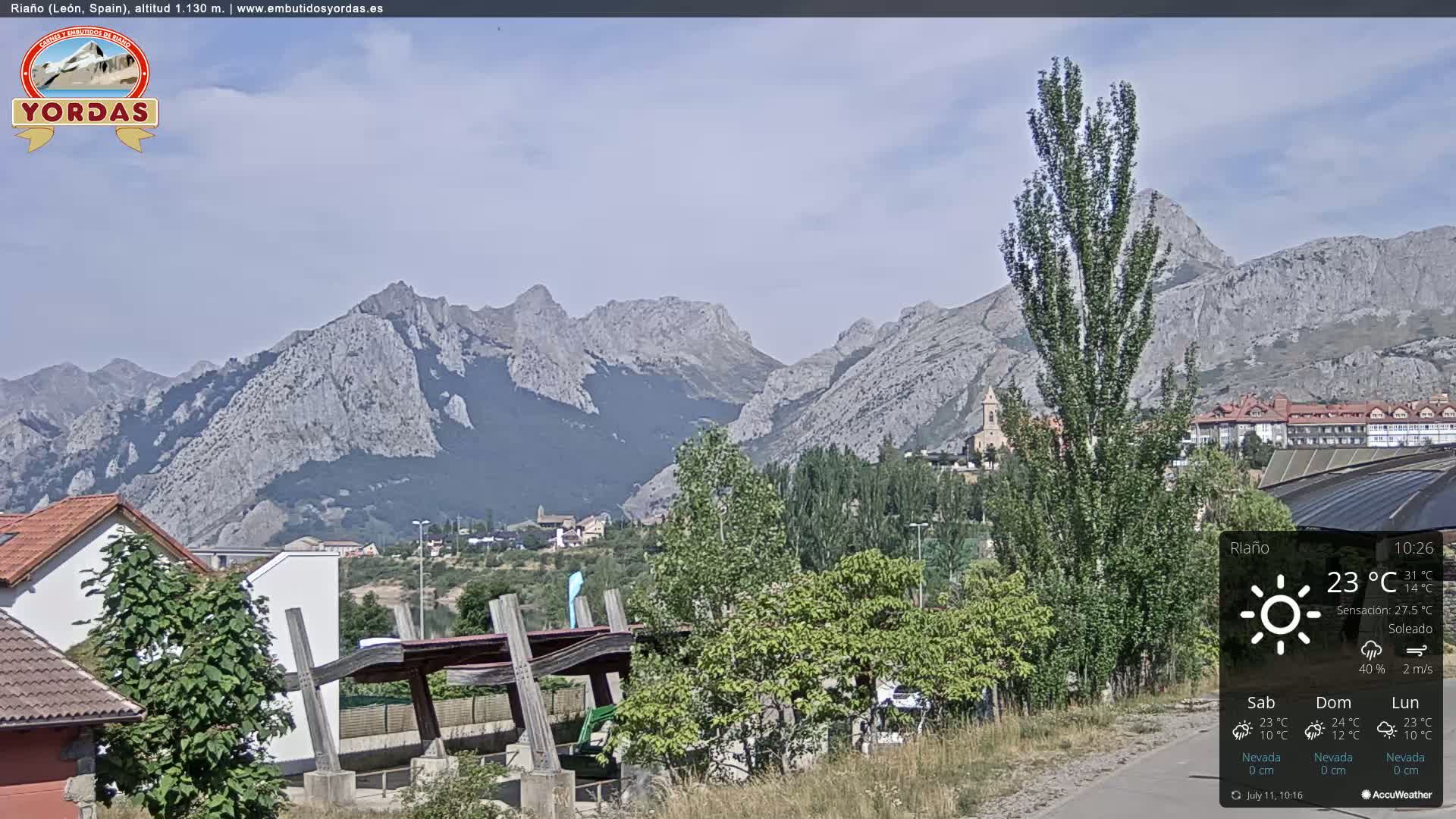 A partly sunny day reveals a mountain range behind a town with green trees and buildings, a wooden structure, and a paved road.