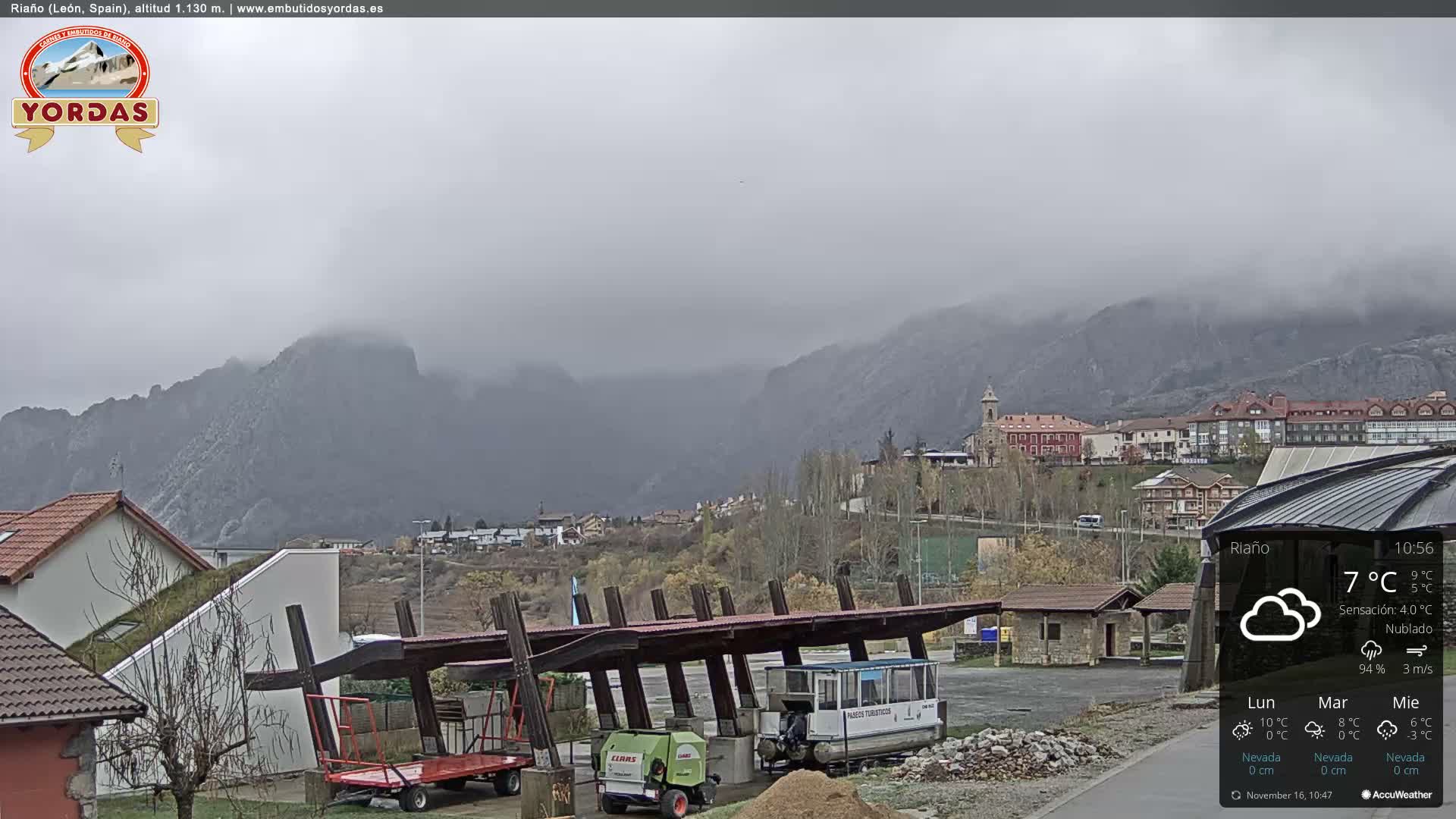 Under a heavily overcast and misty sky, a mountainous landscape with a village featuring a prominent church tower in the valley is seen, while a large covered structure in the foreground shelters a green farm vehicle and a white tourist boat.