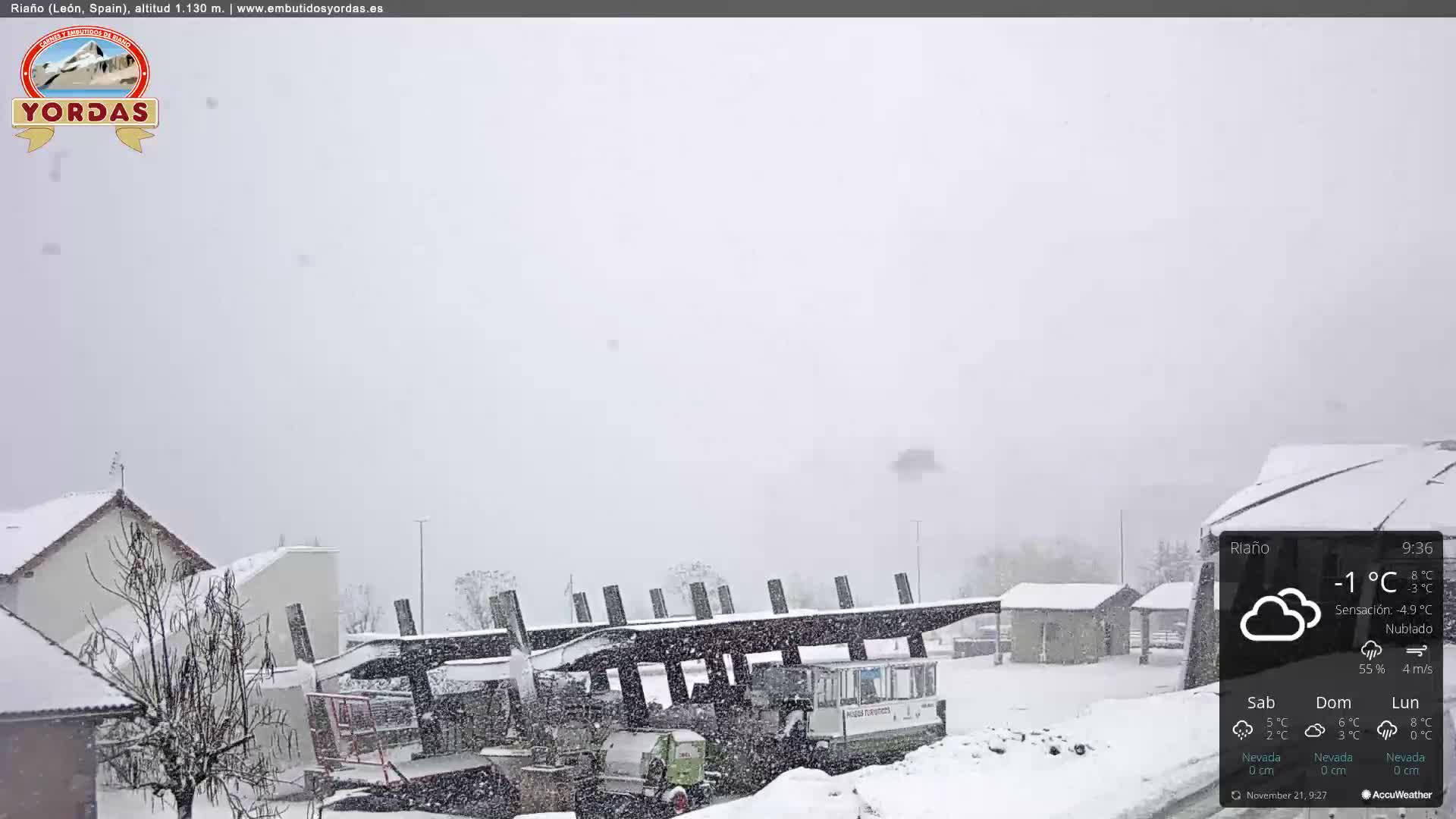 A snowy landscape shows buildings and a large, dark modern structure blanketed in snow, with flakes actively falling under an overcast sky.