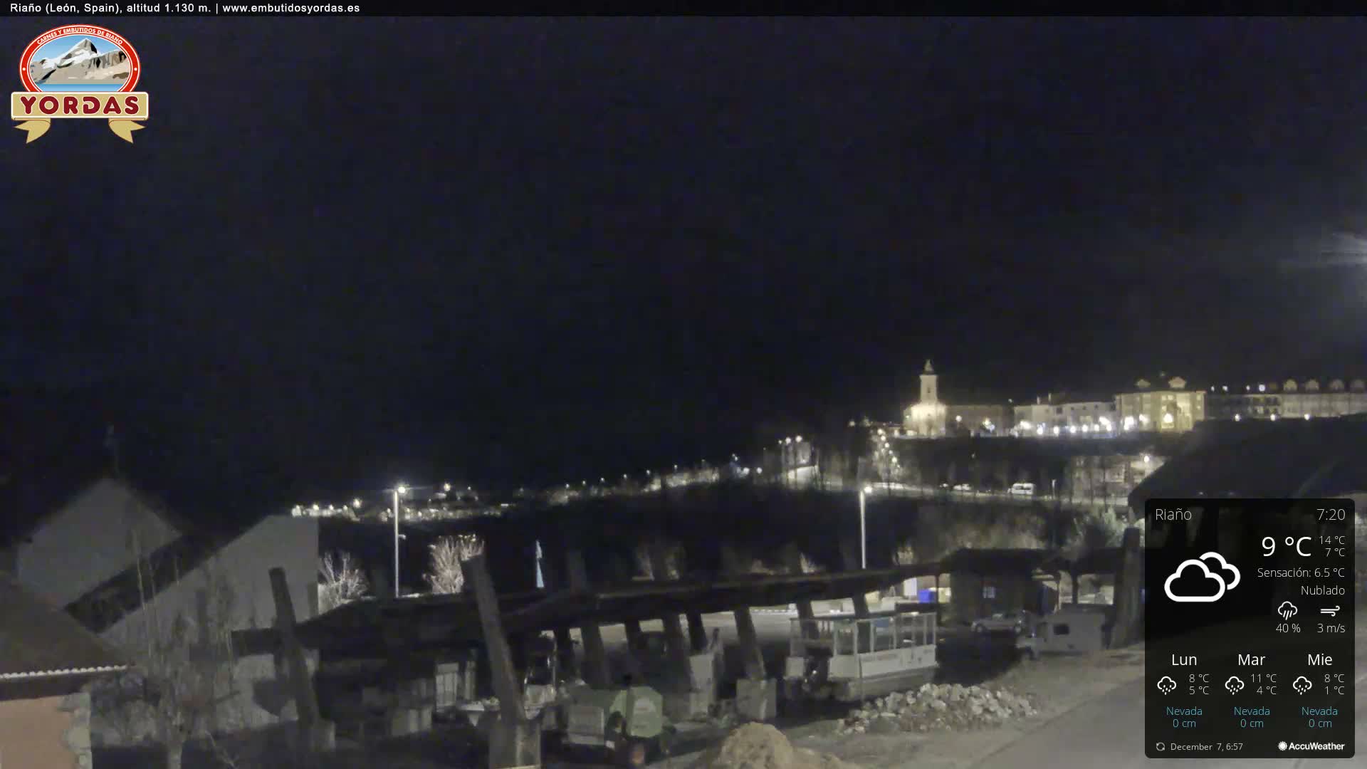 A cloudy night view captures an illuminated town built on a hillside with a prominent church spire in the distance, overlooking a foreground area with bare trees and some light industrial or construction structures.