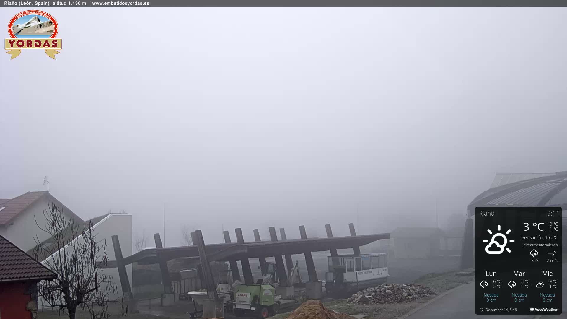 A cloudy night view captures an illuminated town built on a hillside with a prominent church spire in the distance, overlooking a foreground area with bare trees and some light industrial or construction structures.