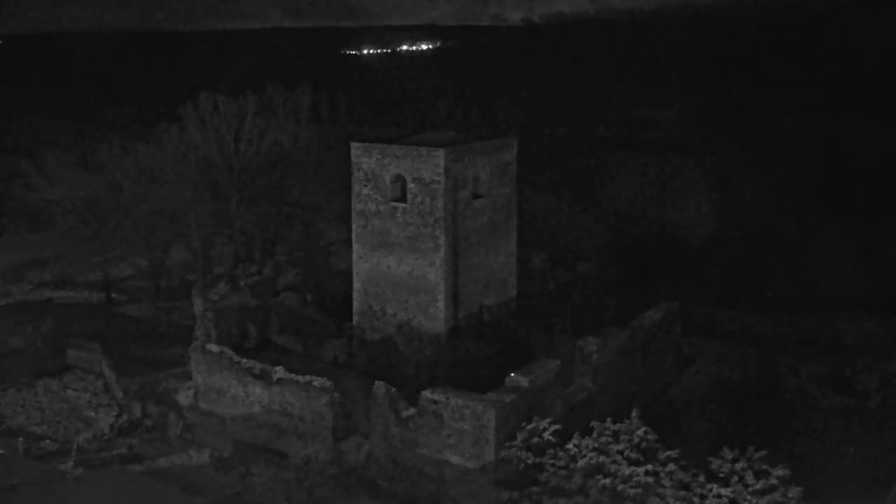 At night, a stone tower stands amidst the ruins of a larger structure, surrounded by dark trees and under a mostly clear sky with distant lights visible on the horizon.