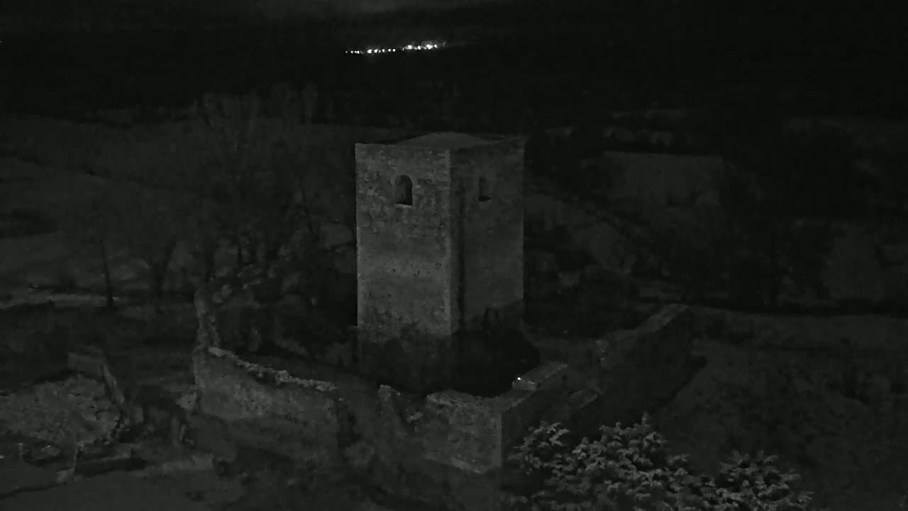 A stone tower, part of a larger ruined structure, stands at night under a dark sky with distant lights visible in the background.